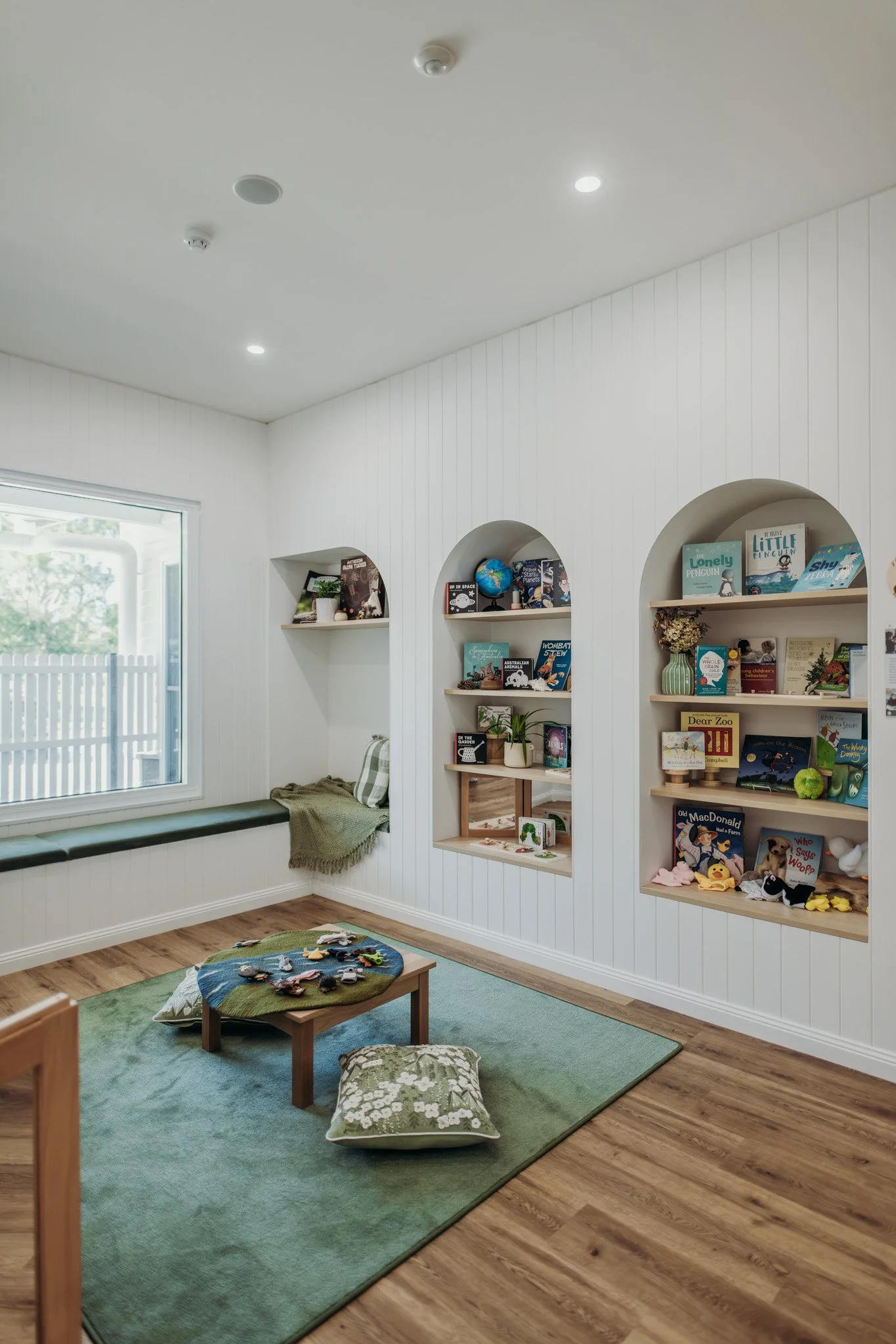A cozy children's reading nook with white paneled walls, built-in bookshelves filled with children's books and toys, a window allowing natural light, a blue and green rug, and floor pillows and a small wooden table with stuffed animals and toys on it