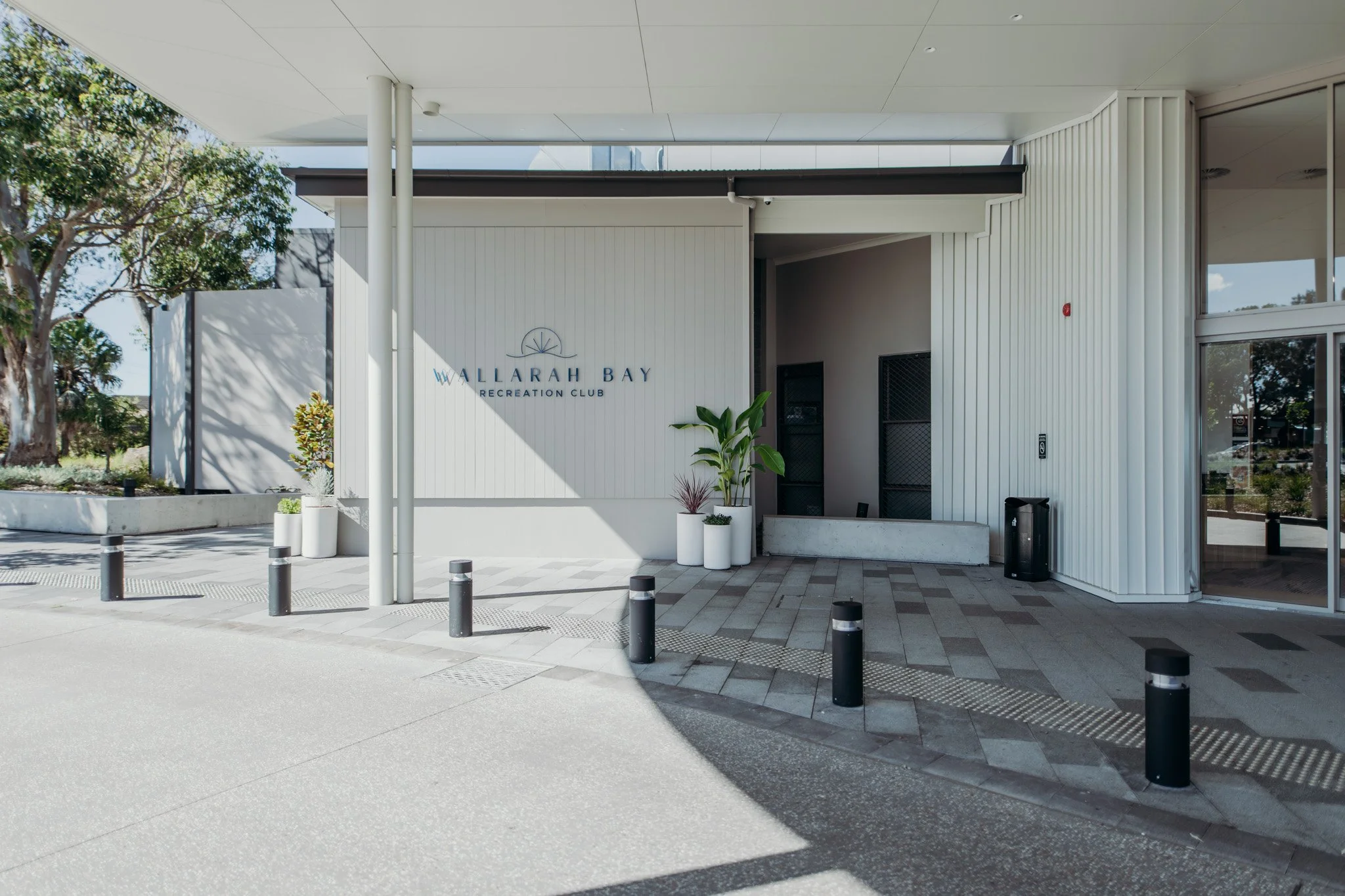 Exterior front of Wallarah Bay Recreation Club building with white siding, large glass windows, potted plants, and a sidewalk with bollards.