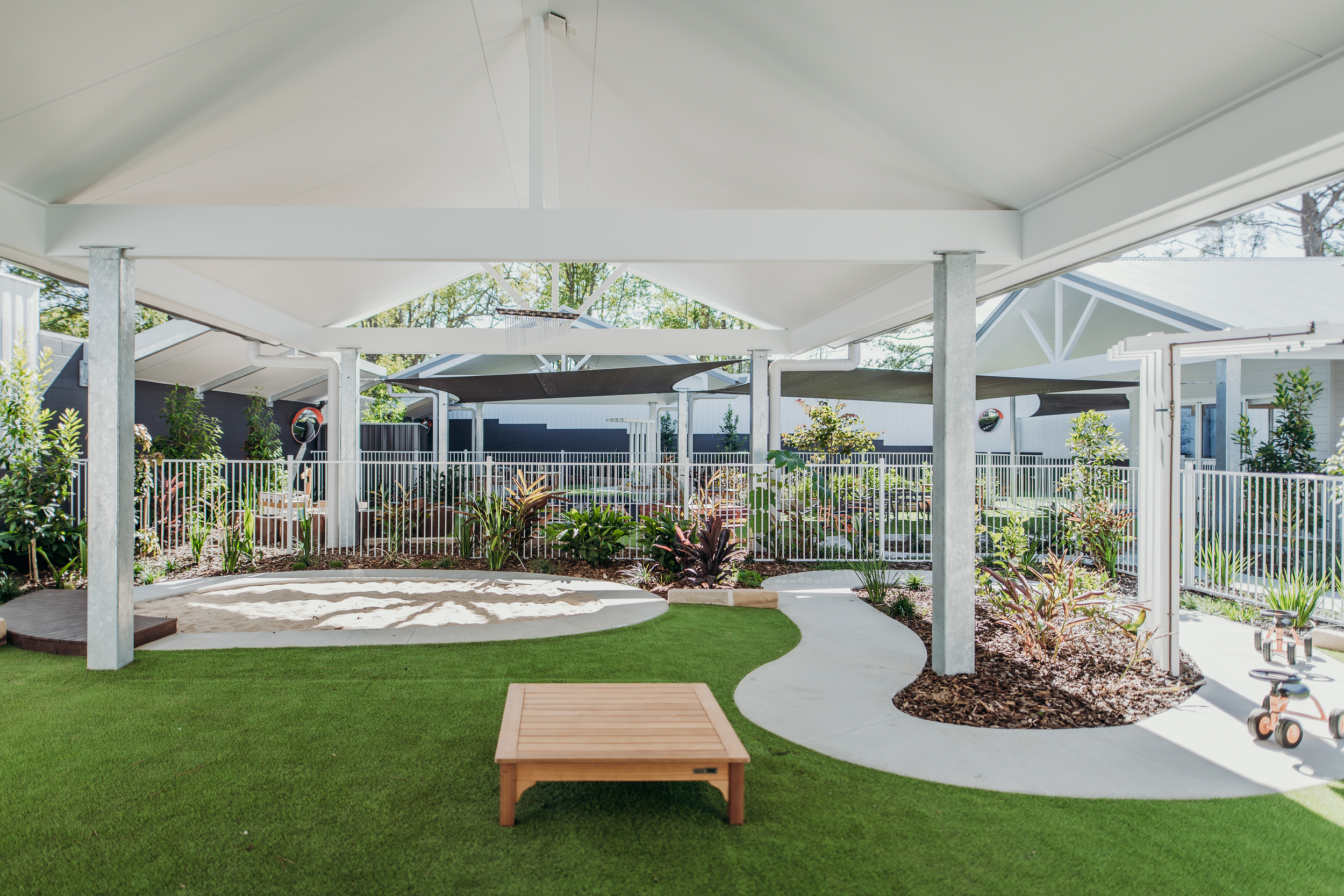 A backyard with a concrete patio, artificial grass, and landscaped garden beds with plants and shrubs, enclosed by a white fence. There is a small wooden table and a sandbox, with shade sails overhead supported by vertical posts. In the background, there are neighboring houses and trees.