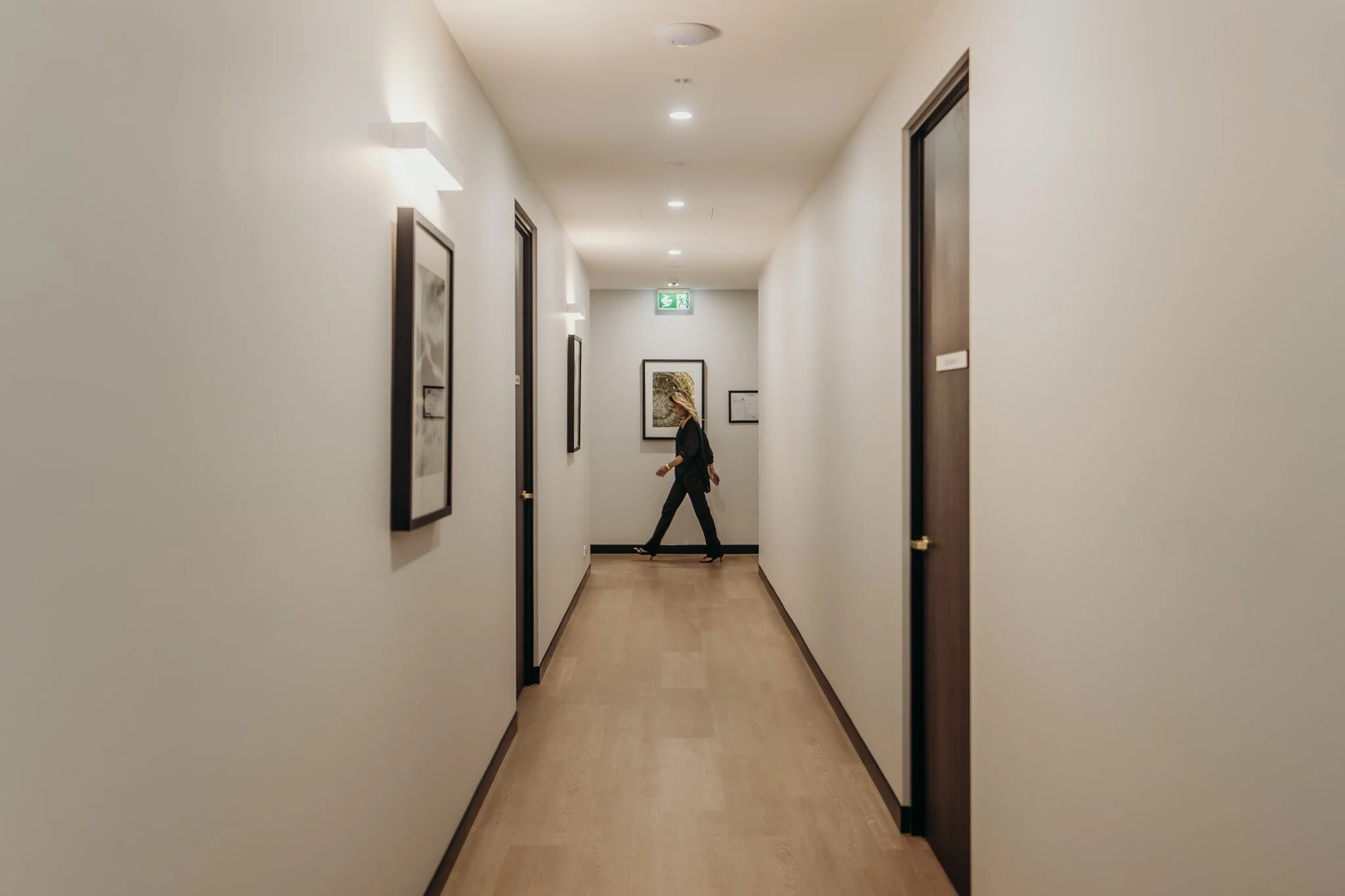 A woman walking down a hallway with framed pictures on the walls and a green exit sign at the end.