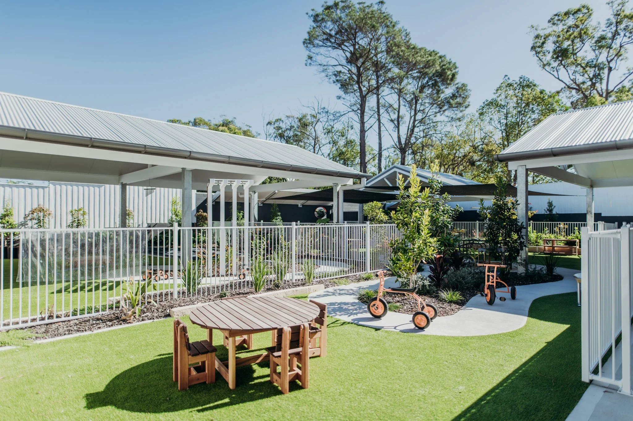 A backyard with a white fence, green artificial grass, a wooden table with chairs, small trees, a toy bicycle, and a modern metal-roofed house against a blue sky with tall trees in the background.