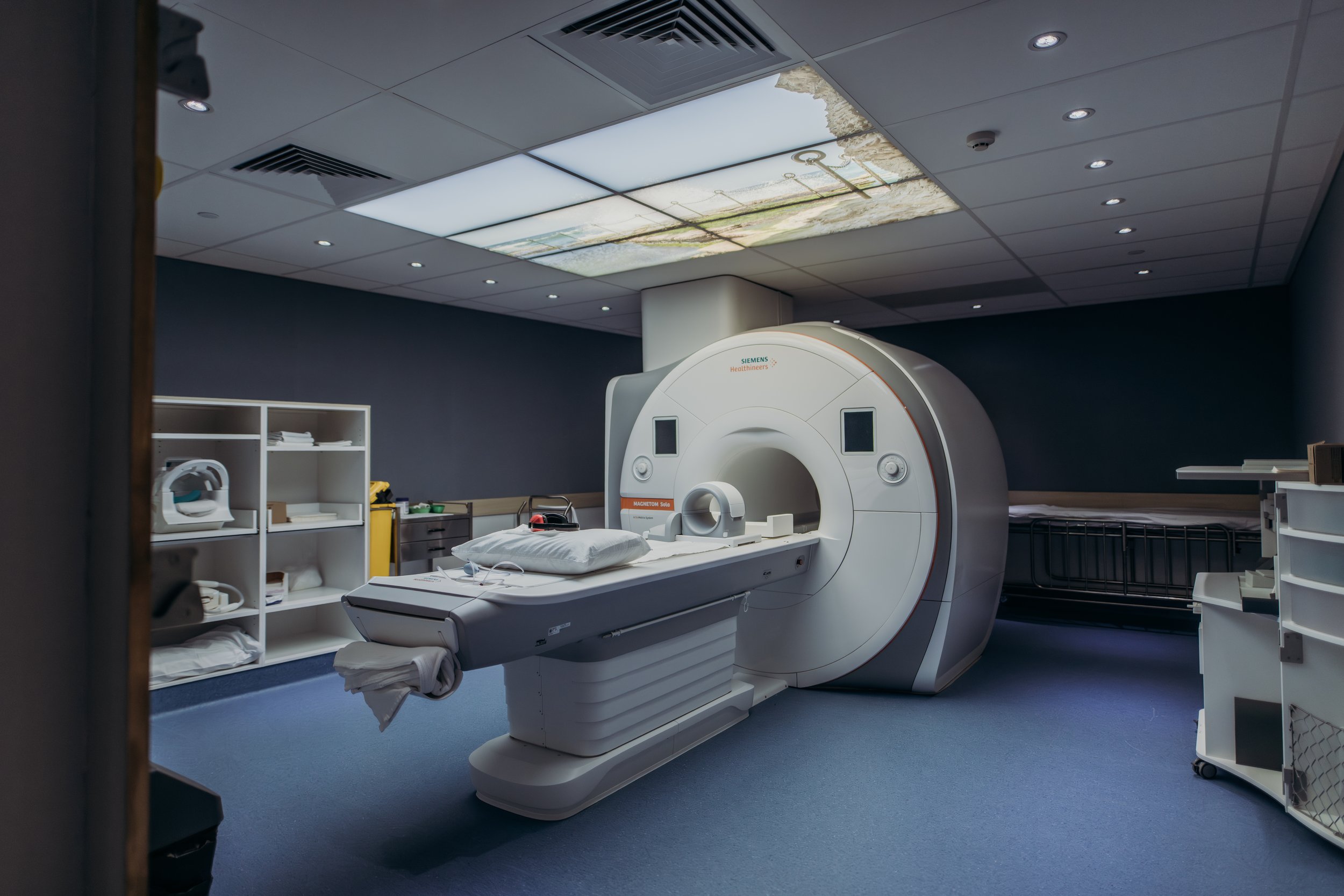Medical room with MRI machine, shelves with supplies, and ceiling with a landscape photograph.