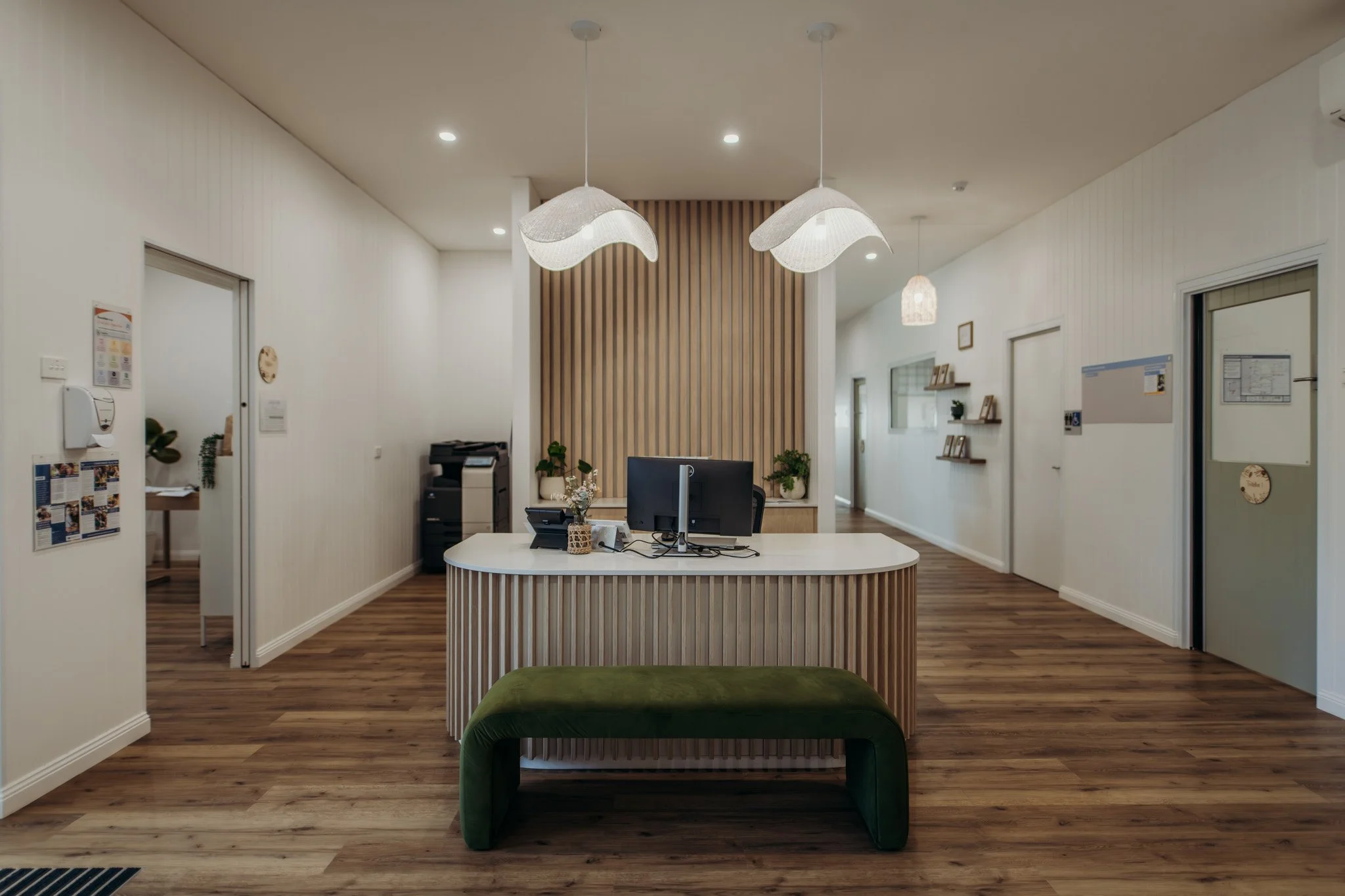 Reception area with a white curved desk, computer, potted plants, and wooden wall paneling behind. Modern pendant lights hanging from the ceiling, white walls, and wood flooring.