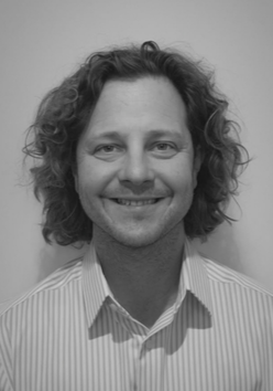 Black and white photo of a man with curly hair, smiling, wearing a striped collared shirt.