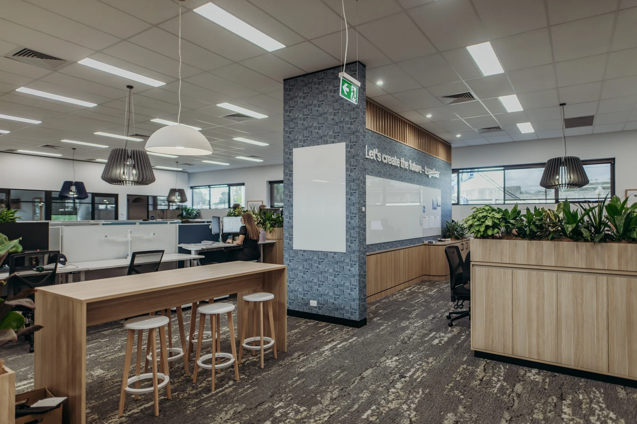 Open office space with desks, chairs, hanging lamps, and green plants. A woman is working at a desk near a wall with quote: "Let's create the future - together."