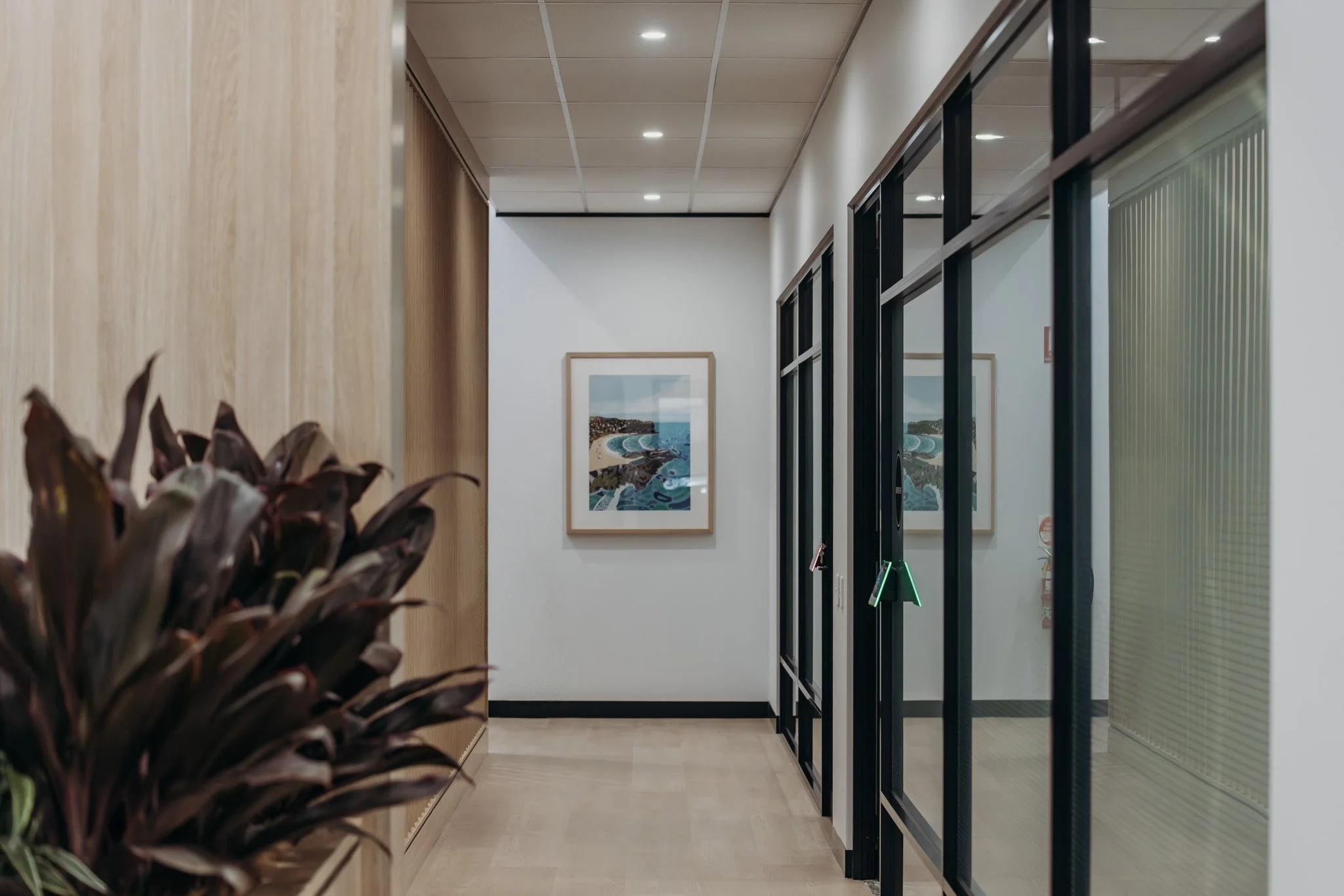 Hallway with framed artwork on the wall, wood paneling on one side, glass doors on the other, and potted plant in the foreground.