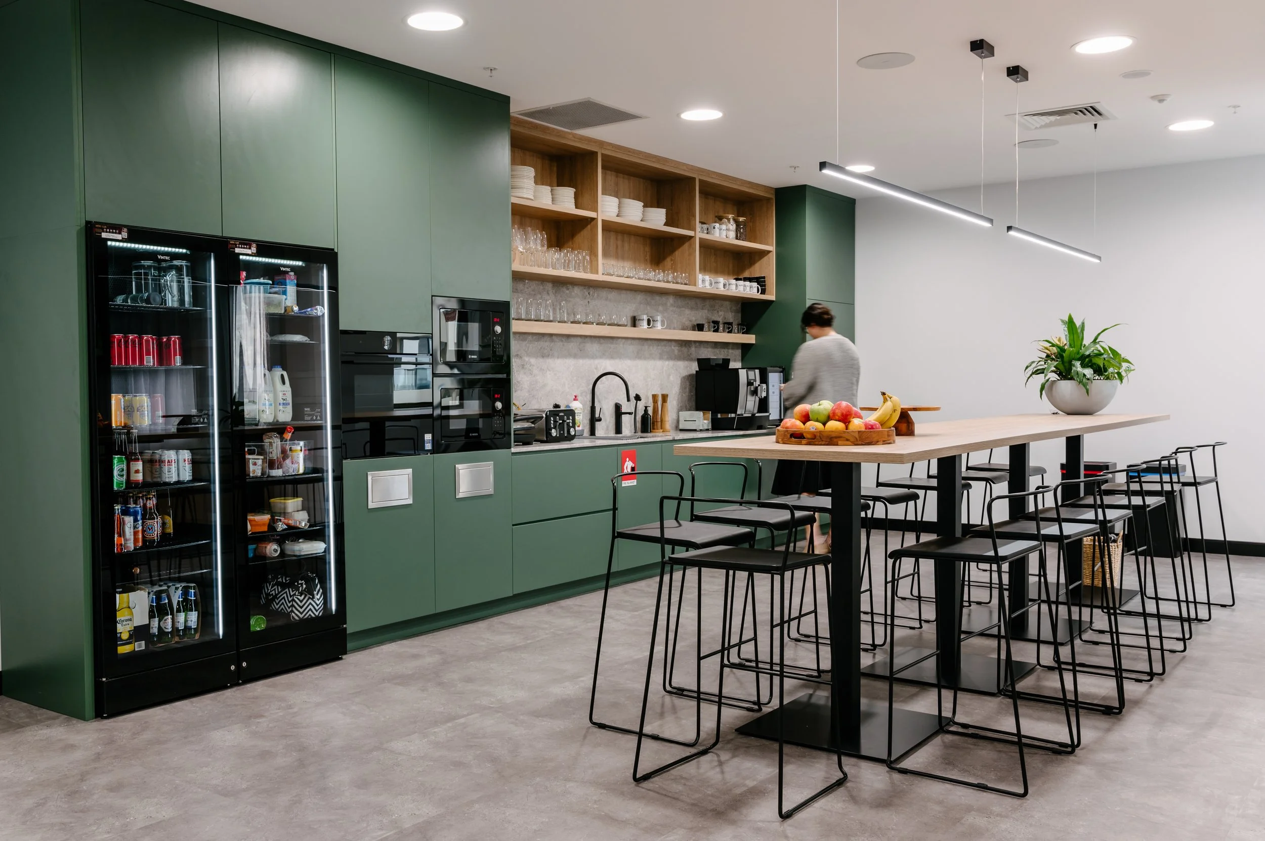 Modern cafe or break room with green cabinetry, open wooden shelves with dishes, a table with chairs, and a large potted plant. A person is working at the counter in the background.