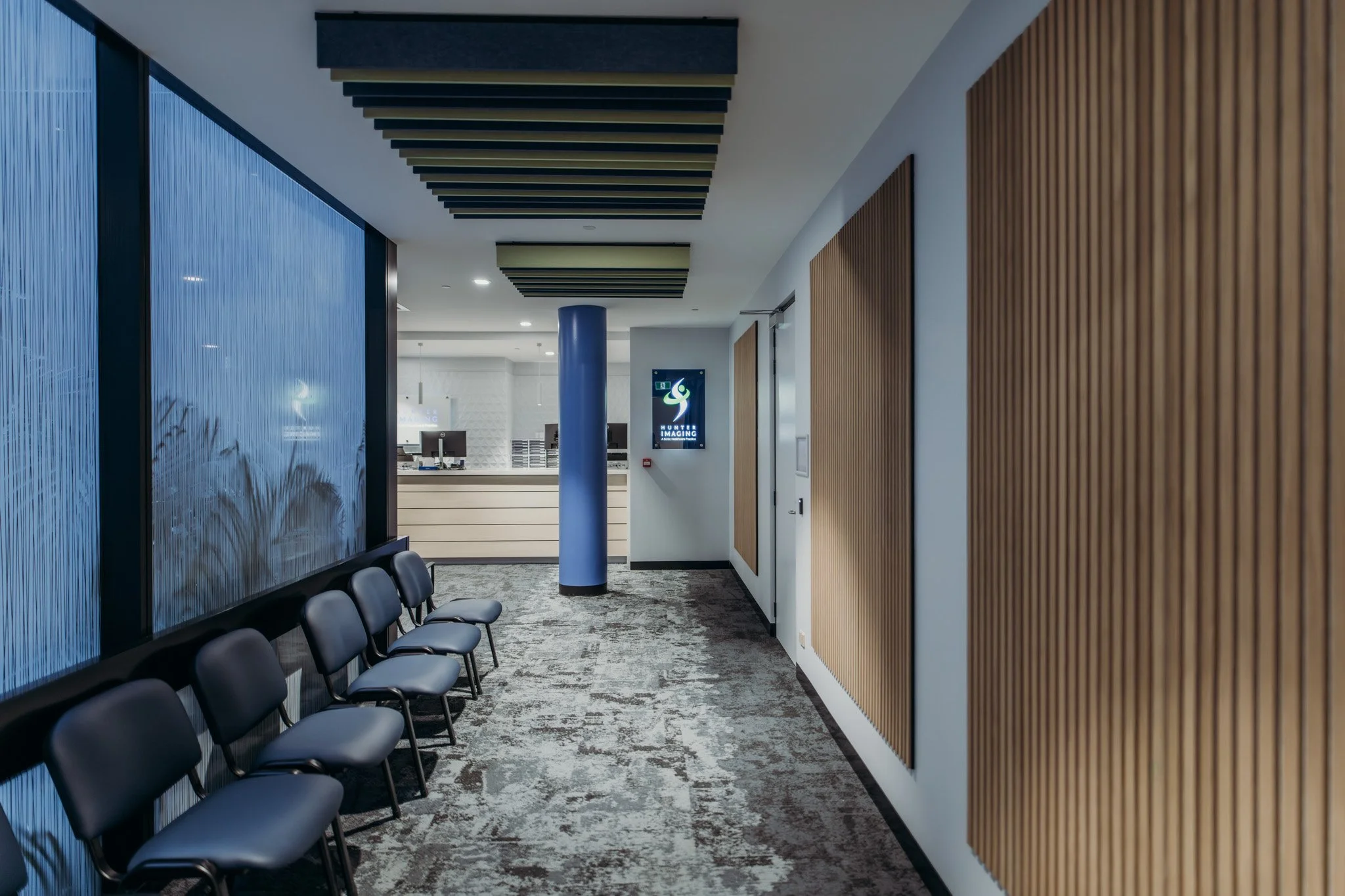 Modern office waiting area with black chairs lined up along a wall, a corridor with wooden wall panels, a blue column, and a reception desk in the background.
