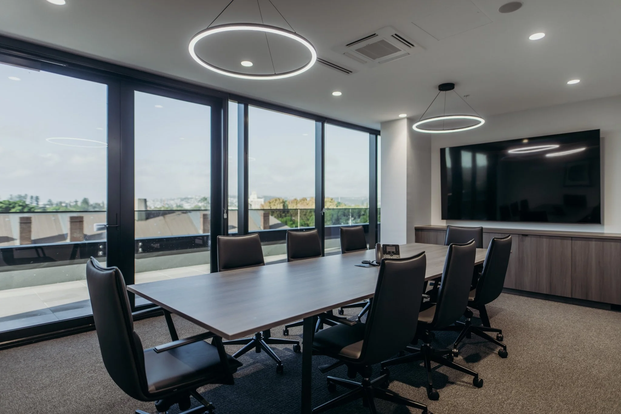 Modern conference room with large windows, black chairs around a table, a large TV screen on the wall, and circular ceiling lights.