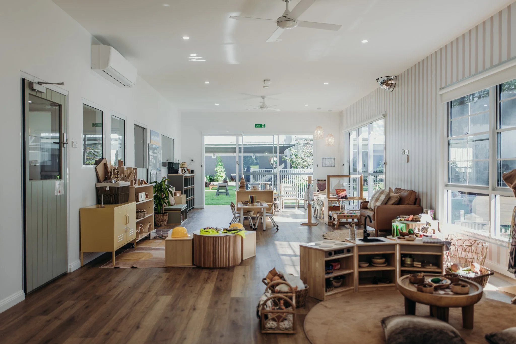 Bright, airy indoor space with wooden floors, white walls, large windows, and a variety of children's furniture and toys. Outside, a small yard with green grass and outdoor furniture is visible.
