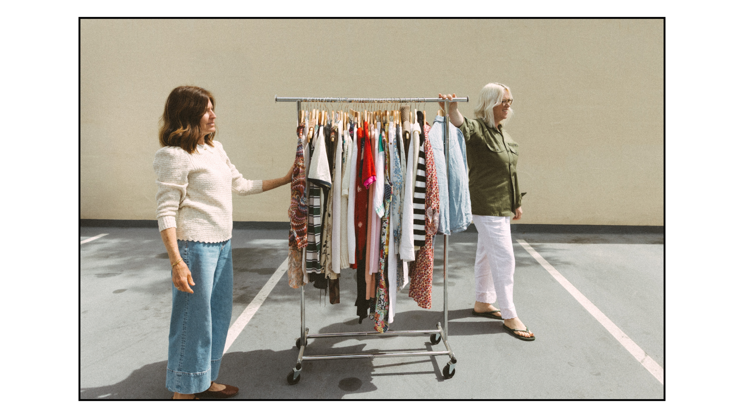 Two women stand on either side of a clothing rack filled with assorted colorful clothes, outdoors in a parking lot.