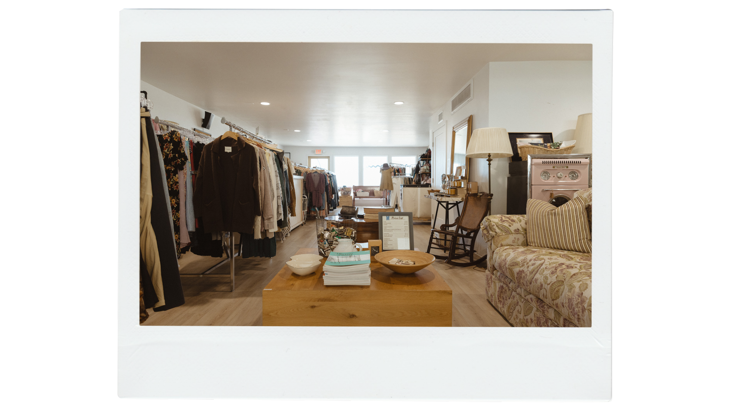 Interior shot of a vintage clothing and home decor store with clothing racks, a cozy sitting area with a floral-patterned sofa, a wooden coffee table with books and bowls, and various decorative items and lamps.