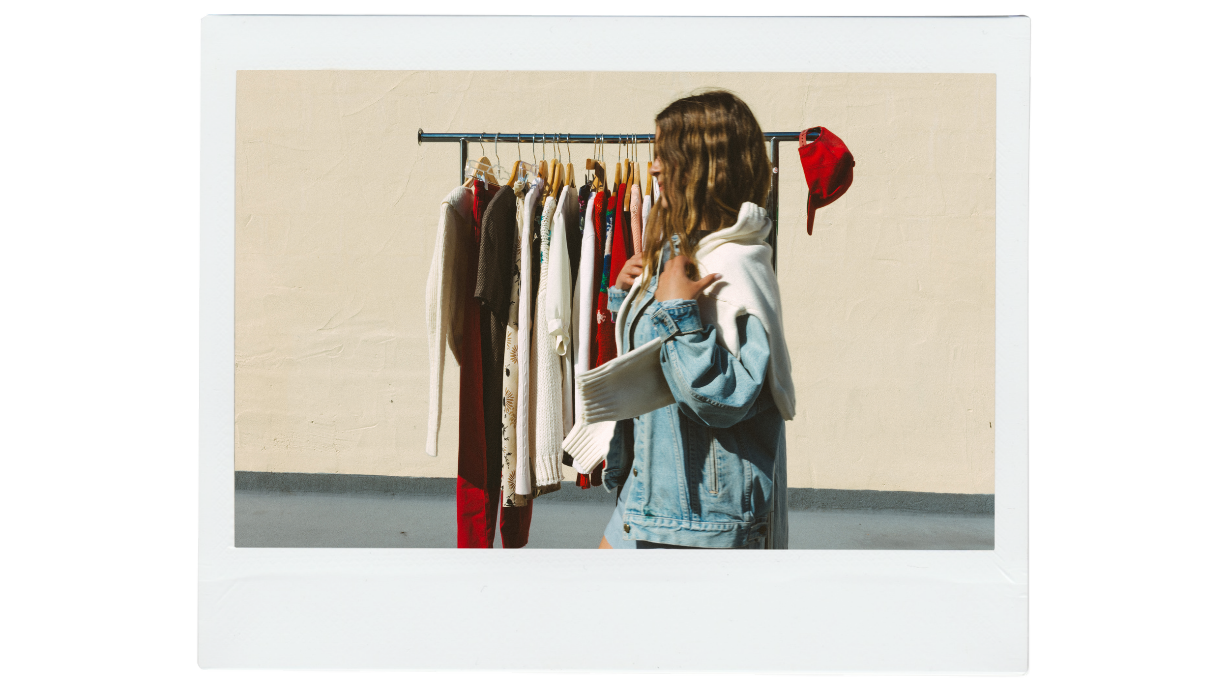 A young woman with long brown hair, wearing a light blue denim jacket and shorts, stands in front of a clothing rack with various garments hanging on wooden hangers. The clothing includes sweaters, dresses, and shirts in different colors and patterns. She is outdoors against a plain off-white wall, and a red baseball cap hangs on the end of the clothing rack.