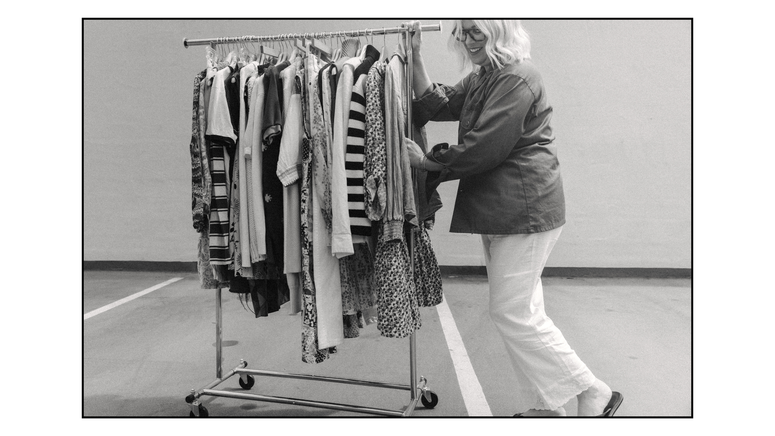 A woman with blonde hair and glasses shopping for clothes at a clothing rack in a parking lot.