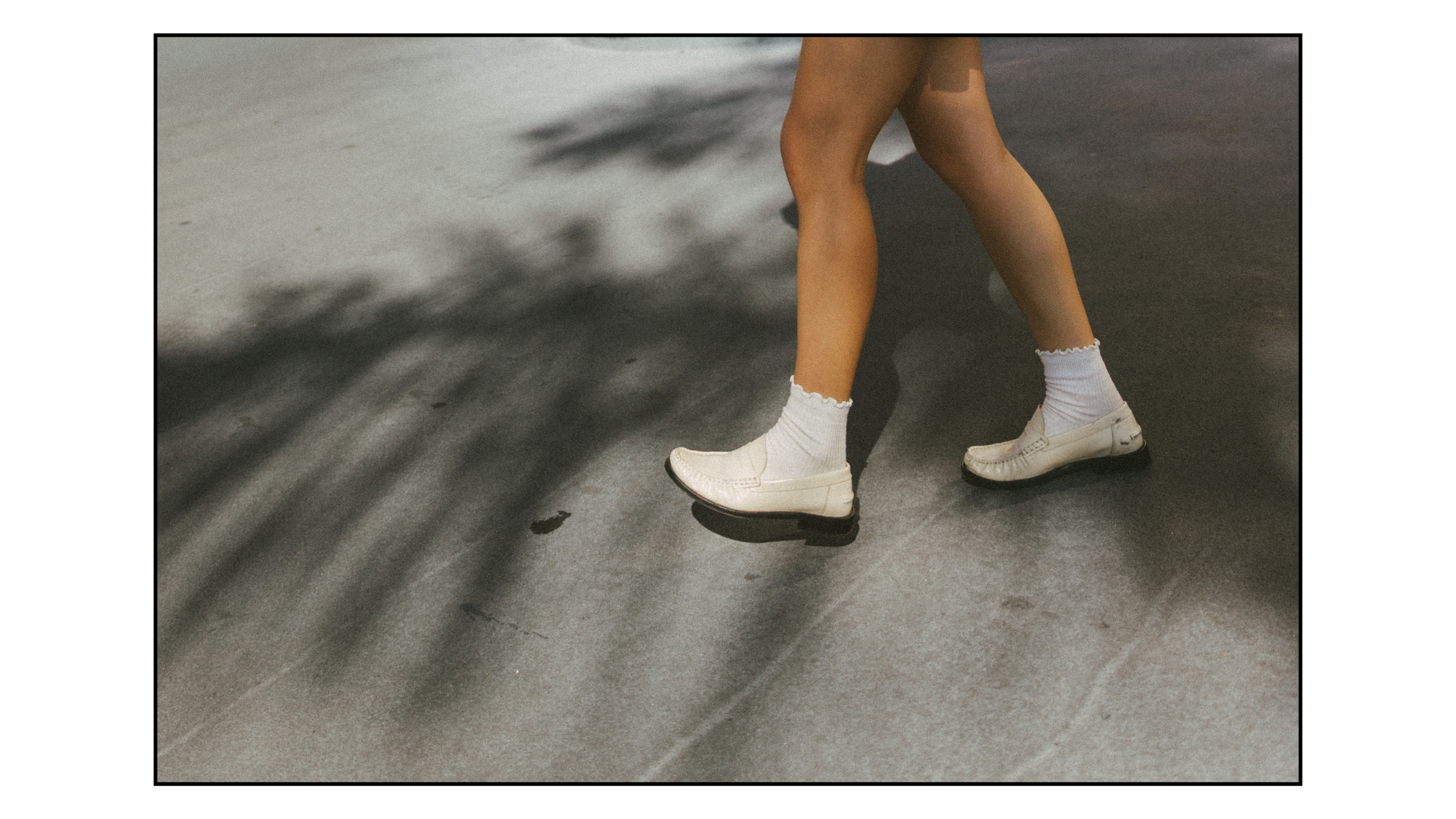 Close-up of a person's legs wearing white loafers and white socks, walking on a dark paved surface with shadows.
