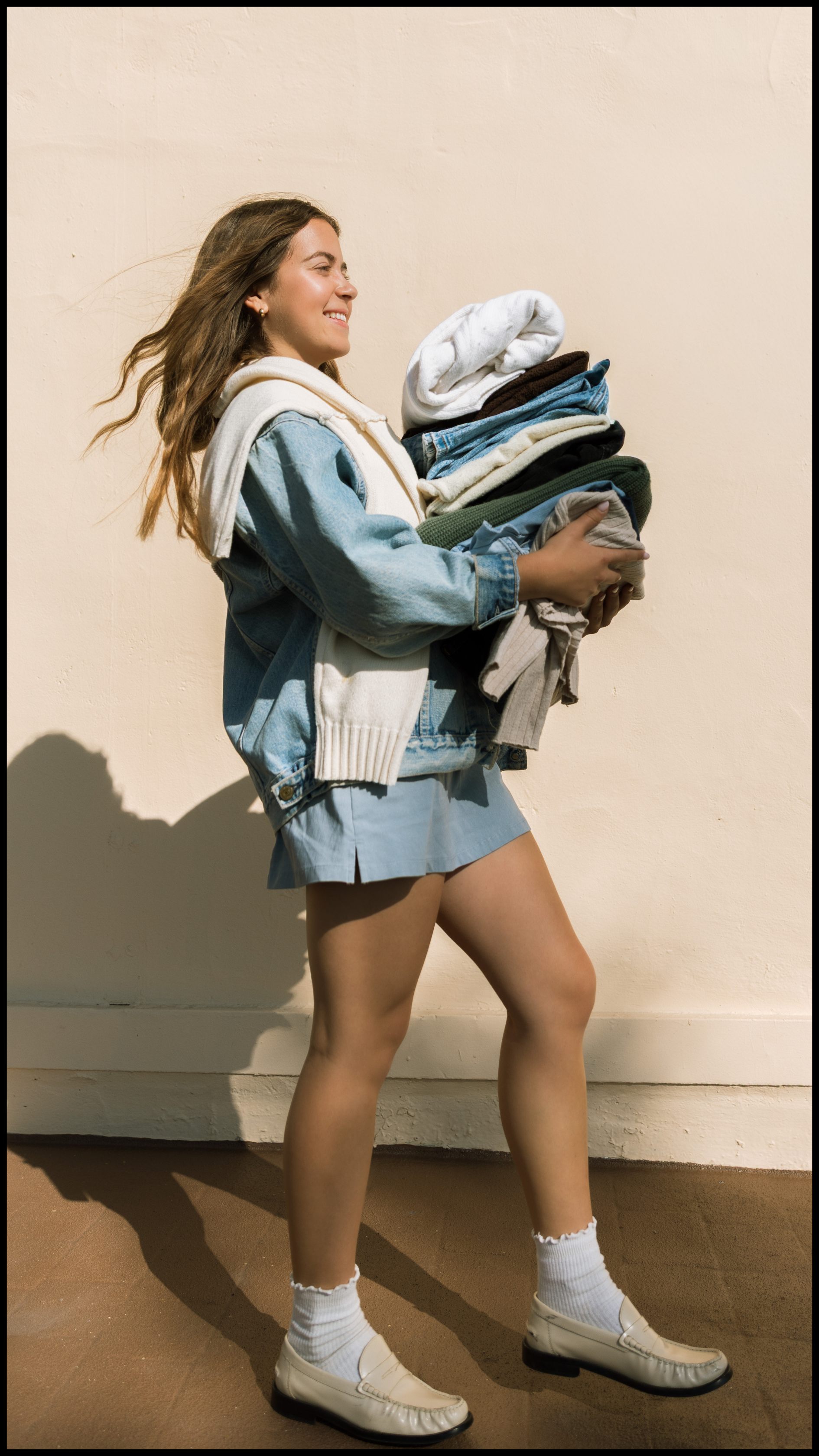 A young woman with long brown hair smiling while walking, carrying a large pile of folded clothes. She is wearing a light-colored hoodie draped over her shoulders, a denim jacket, a short skirt, white socks, and beige loafers, standing against a beige wall with sunlight casting shadows.