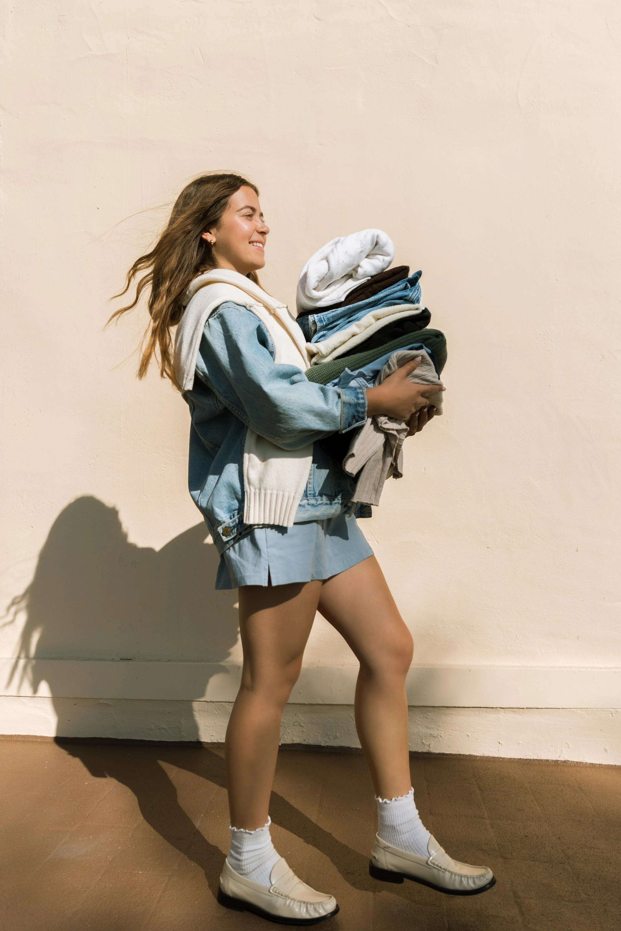 A young woman with long brown hair smiling and walking while holding a pile of folded clothes against a plain beige wall. She is dressed in a denim jacket, gray skirt, white sneakers, and white socks.
