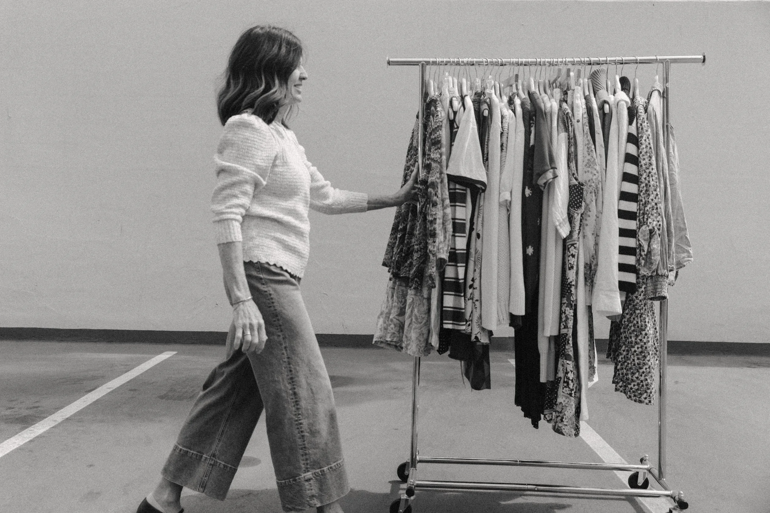 A woman with wavy hair walking past a clothing rack with various garments in an indoor setting, possibly a garage or parking lot.