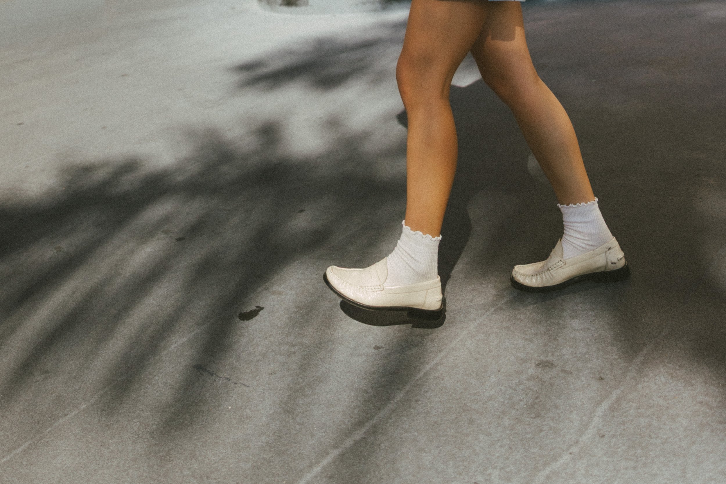 Close-up of a person's lower legs and feet, wearing white sneakers and white socks, standing on a dark concrete or asphalt surface with shadows of nearby objects.