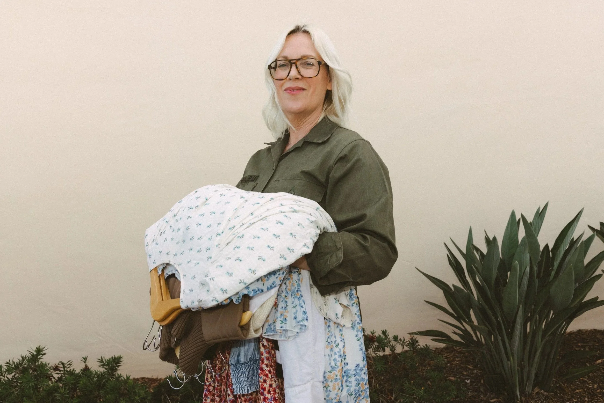 A woman with blonde hair, glasses, and a gray jacket holding a baby wrapped in a white blanket with blue patterns, standing next to a large green plant against a plain beige wall.