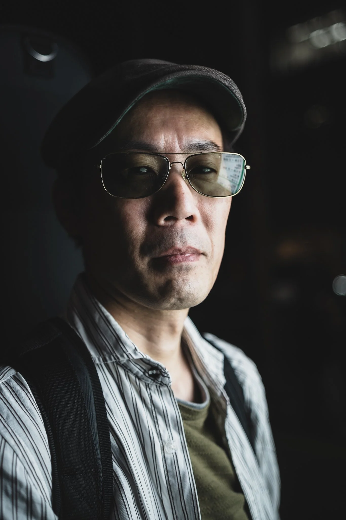 キャップをかぶりサングラスをかけた男性の顔のアップ写真。暗い背景と自然な表情。