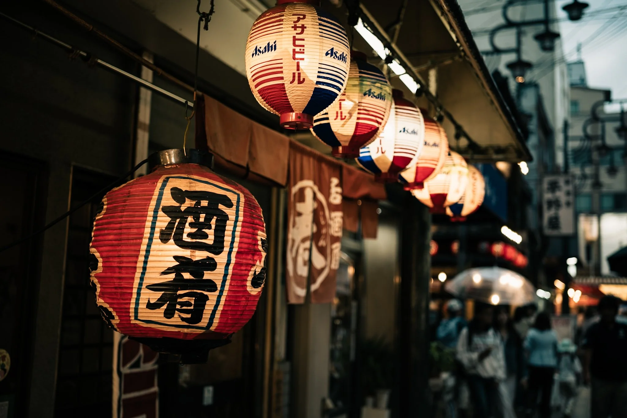 提灯に日本語やブランド名が書かれている屋台の飾り。夜の街並みの中、店舗や商店の外に並んでいる。」}》}