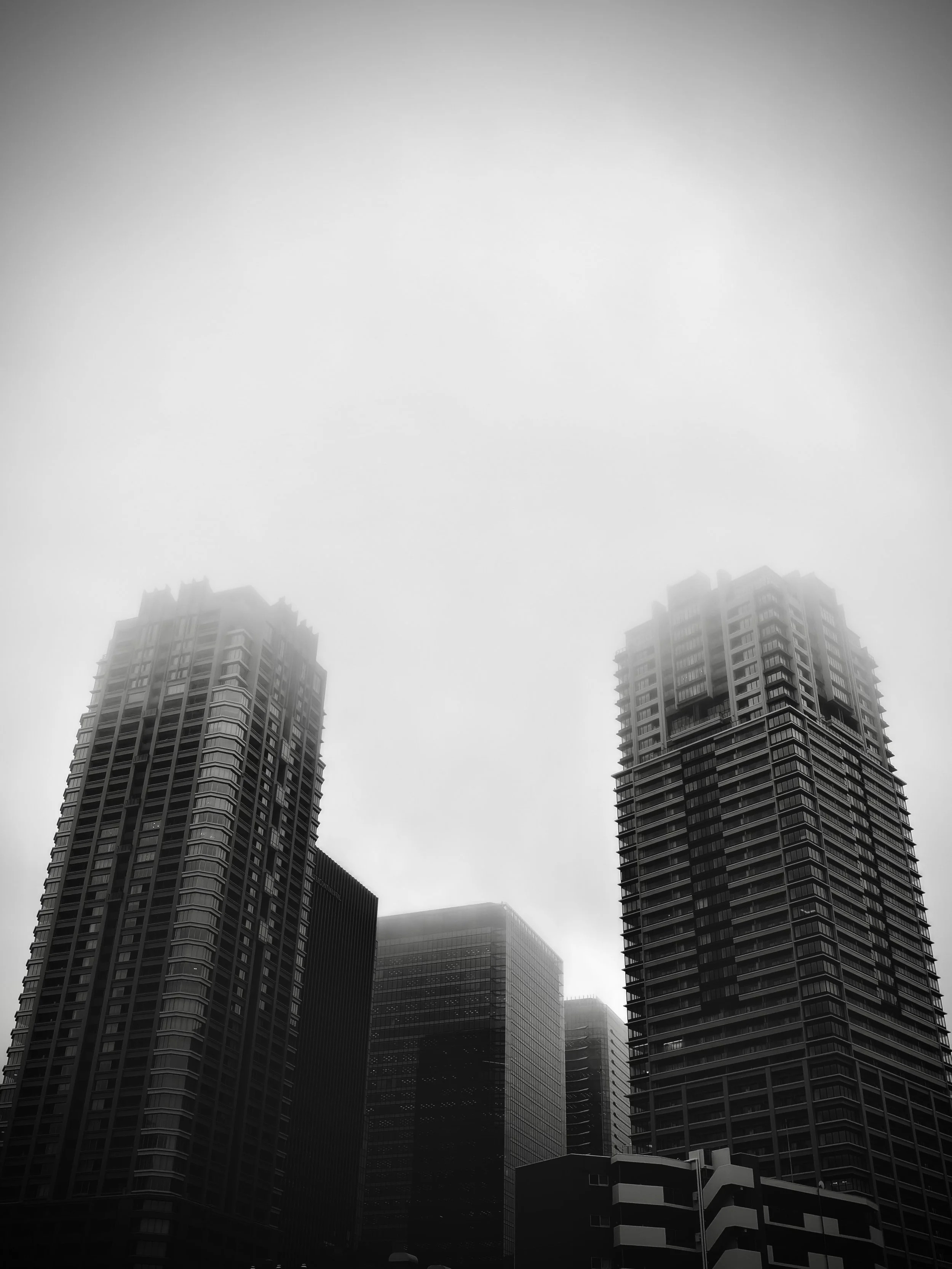 高層ビルが密集した都市の風景。曇り空の下、ビルの上部が霧に包まれている。黒と白のモノクロ写真。