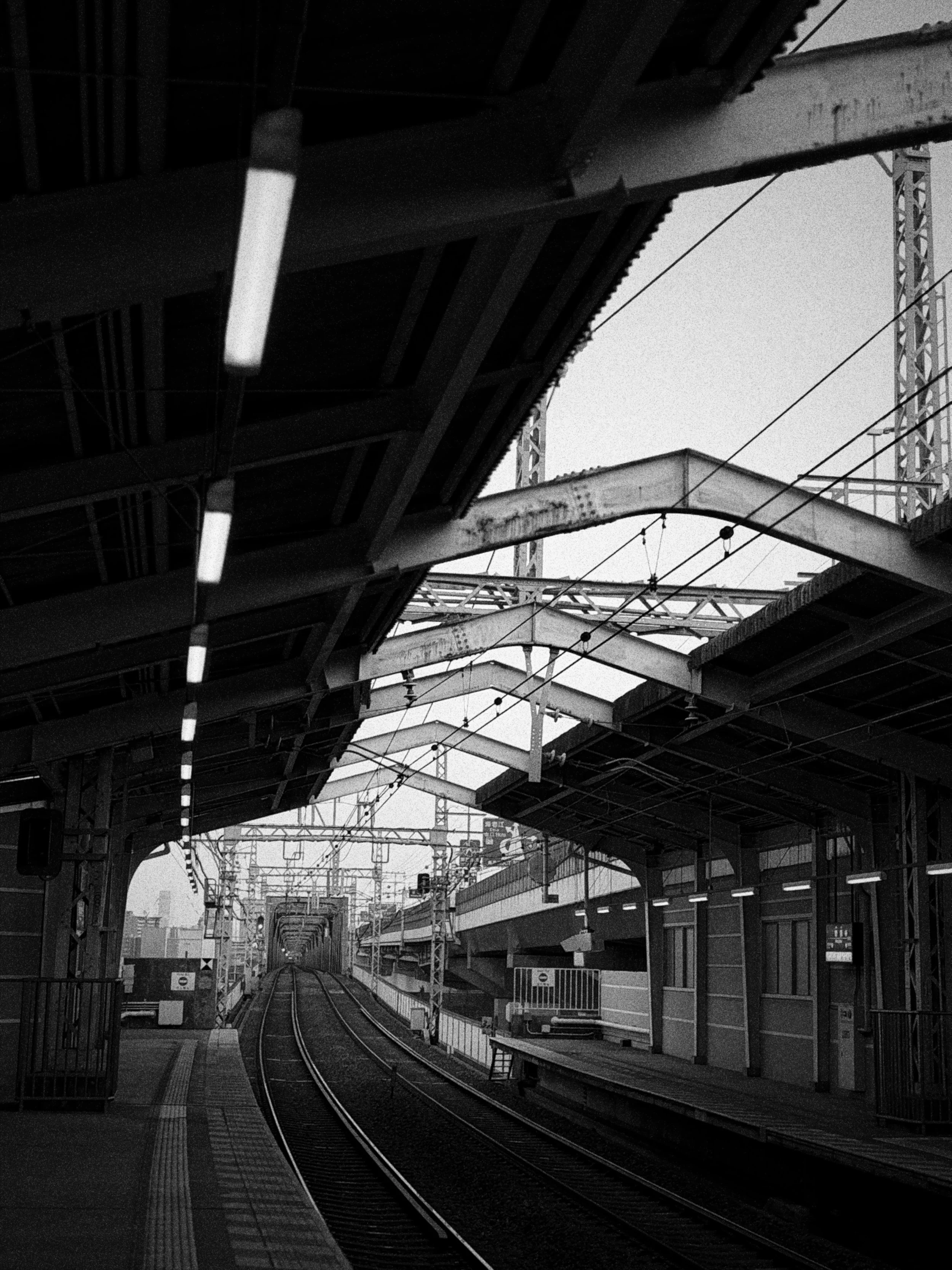 都会の鉄道駅のホームと線路。天井には電灯が並び、天候は曇り。
