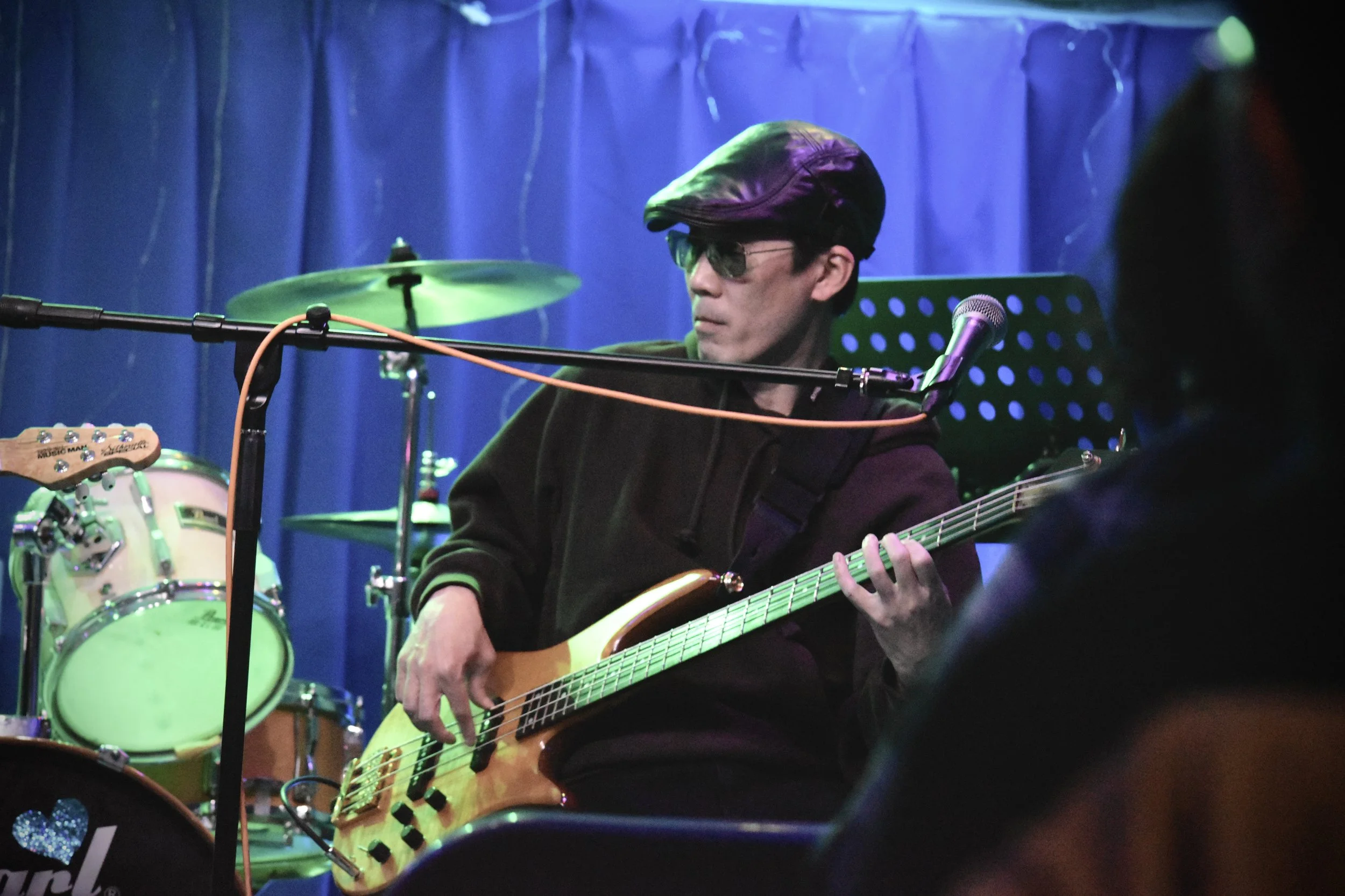 サングラスをかけた男性がギターを弾いているライブ演奏のシーン。背景は青いカーテンとドラムセットが見える。