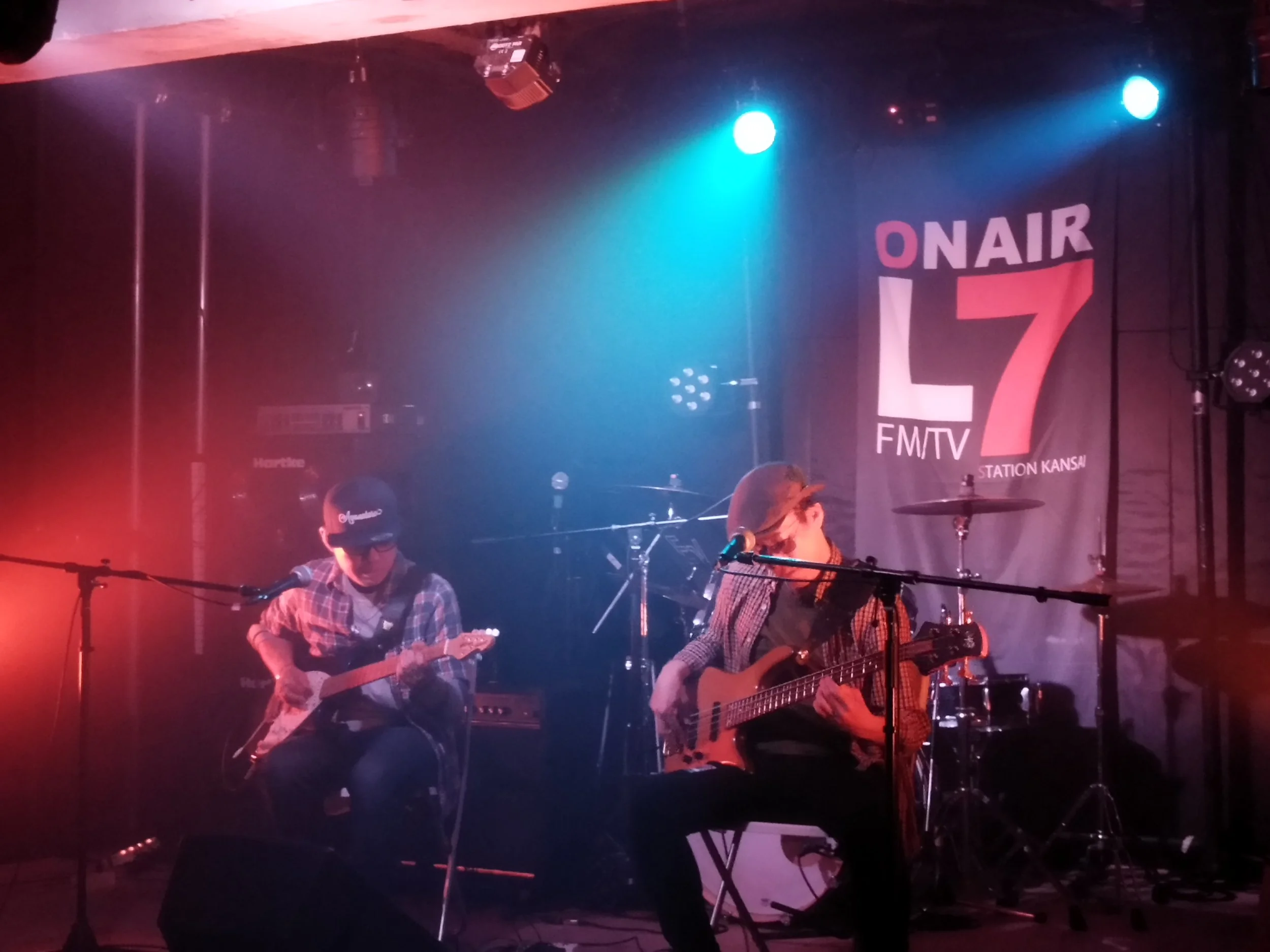 音楽ライブのステージ上でギターとドラムを演奏するミュージシャンたちと「ON AIR 7 FMTV STATION KANSAI」のサイン