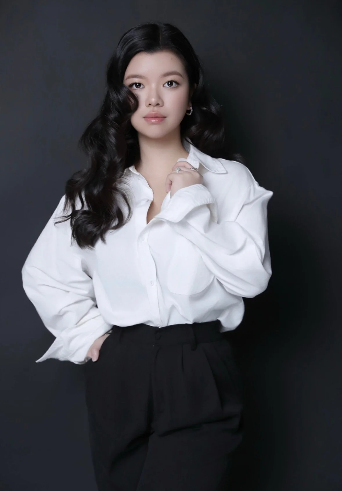 A woman with long, black, wavy hair wearing a white button-up shirt and black pants against a dark backdrop.