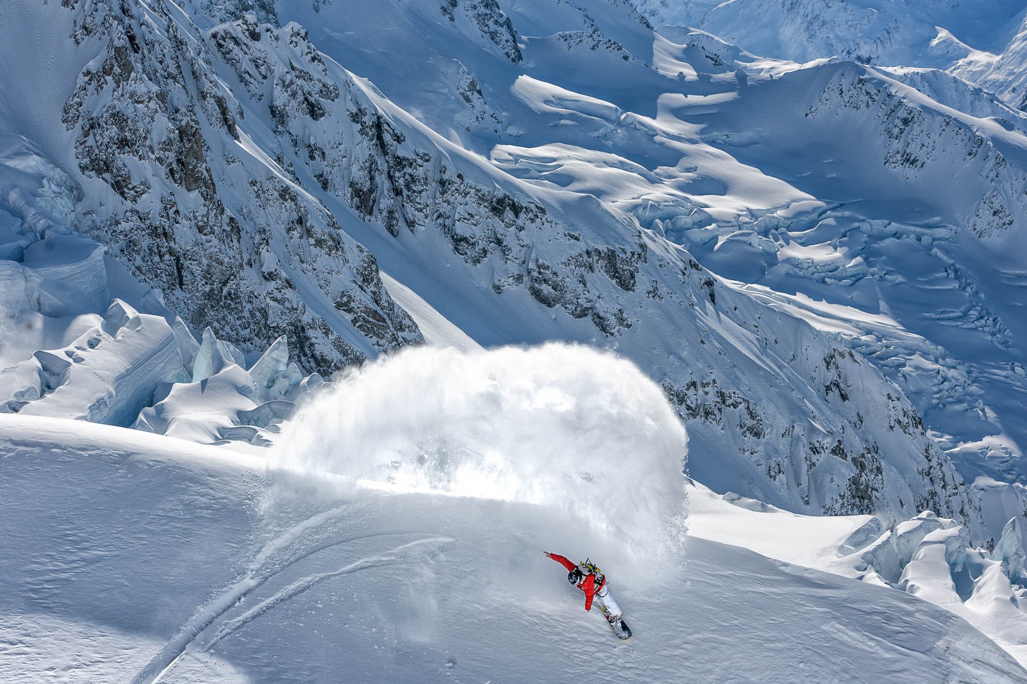 A skier in red gear carving through deep snow on a mountainous, snow-covered slope with steep rocky cliffs in the background.