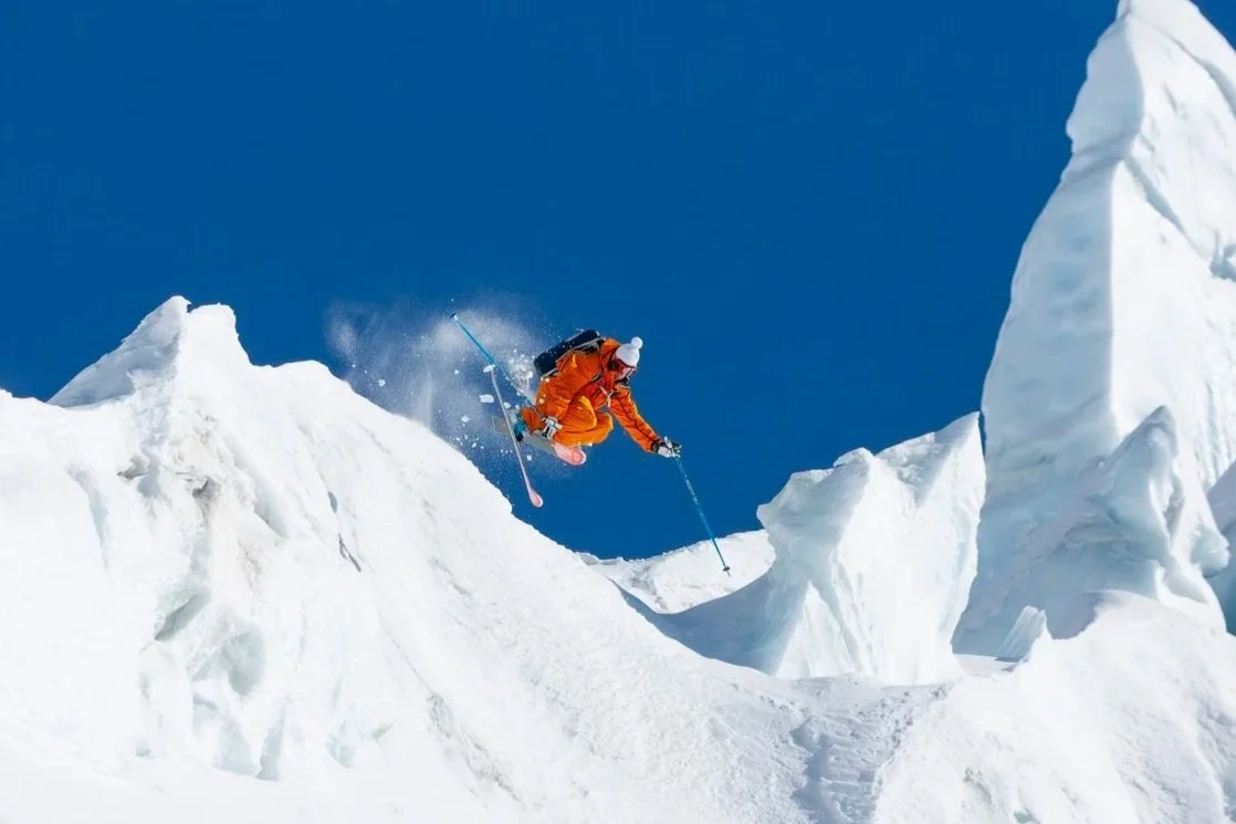 A person in orange snow gear skiing downhill on a snowy mountain with large snow formations and a clear blue sky.