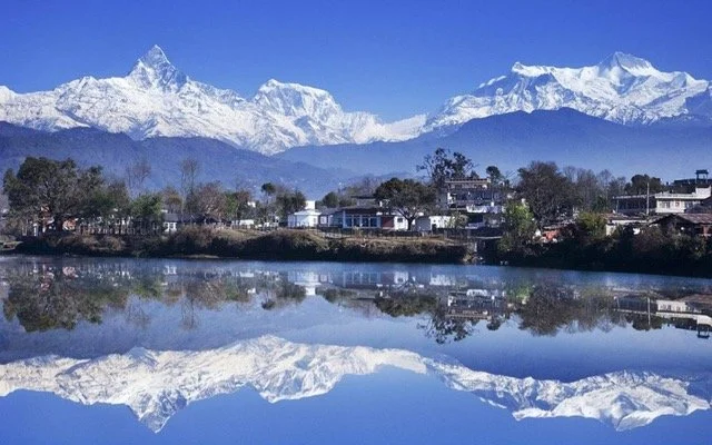Snow-capped mountains and a clear lake reflecting the mountains and trees with a small town in the foreground under a blue sky.