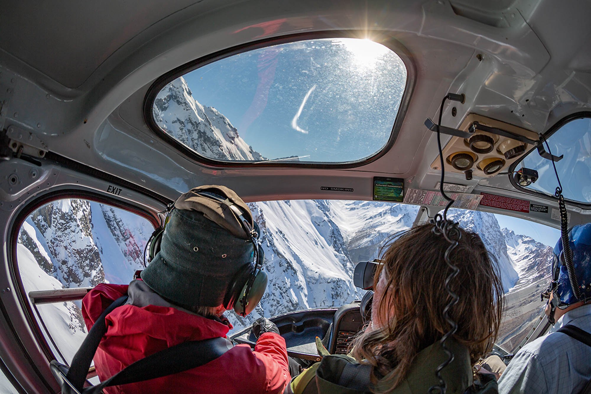 View through helicopter window showing snow-covered mountains and blue sky with sunlight.