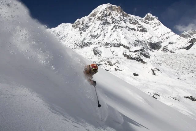 Skier in a helmet skiing down a snowy slope with a snowy mountain in the background.