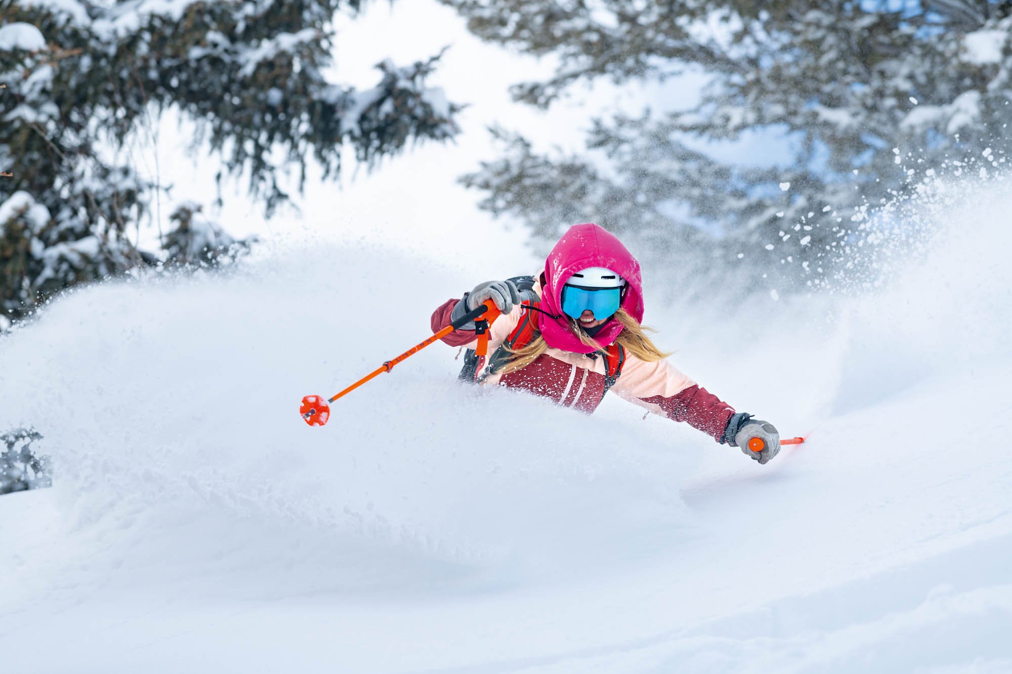 A person wearing a pink winter hat, goggles, and winter gear skiing down a snowy slope, kicking up snow behind them, surrounded by snow-covered trees.