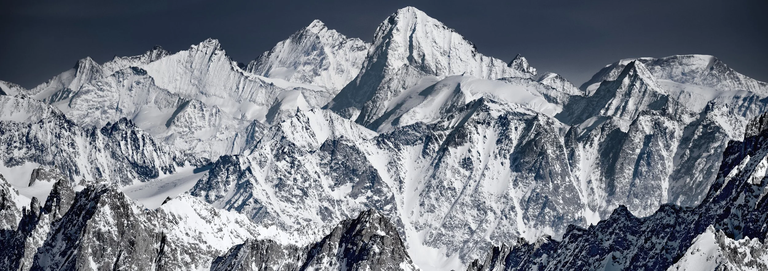 Snow-capped mountain peaks in a rugged mountain range under a dark sky.