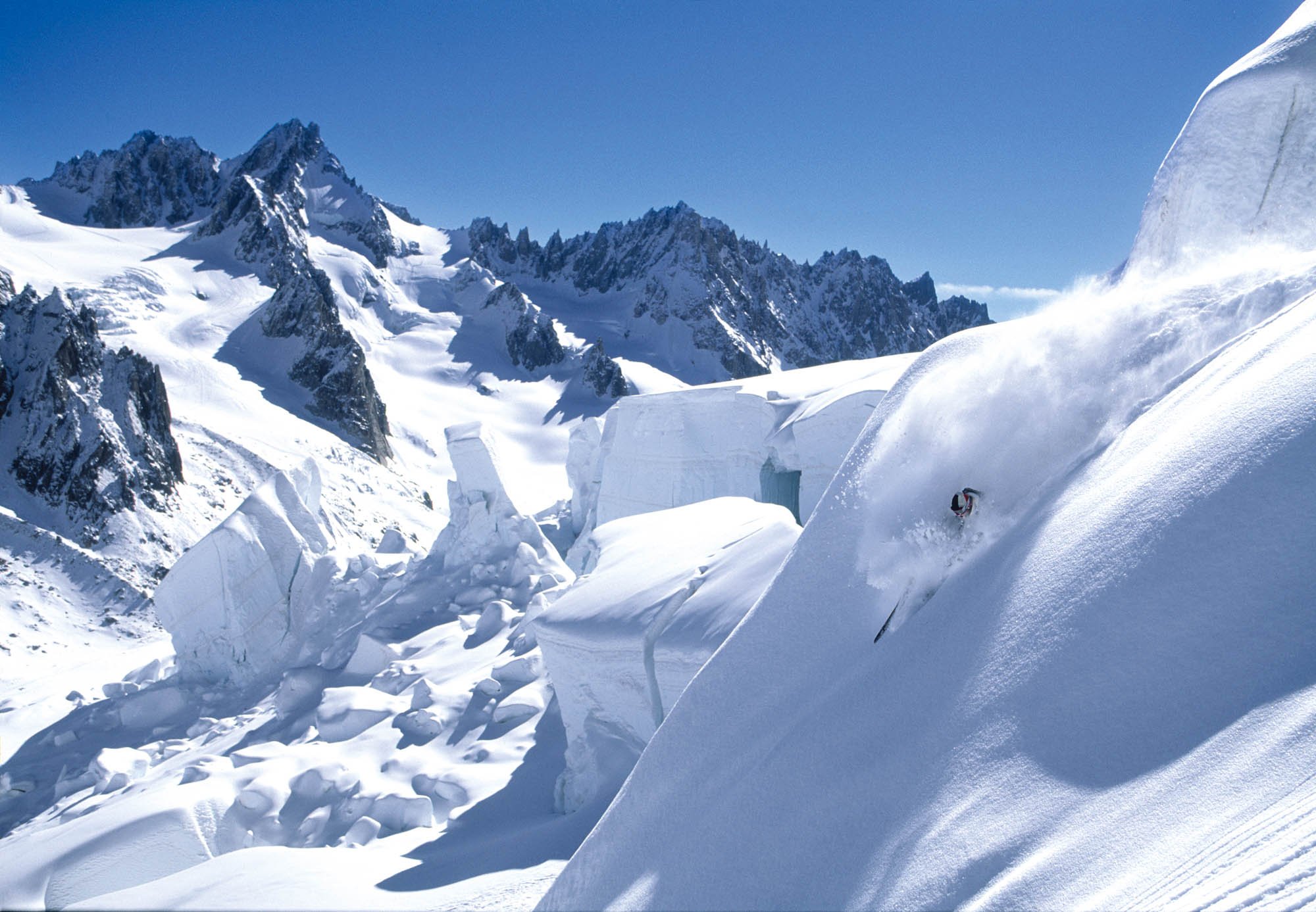Snow-covered mountain landscape with jagged peaks under clear blue sky, and an ice formation in the foreground.