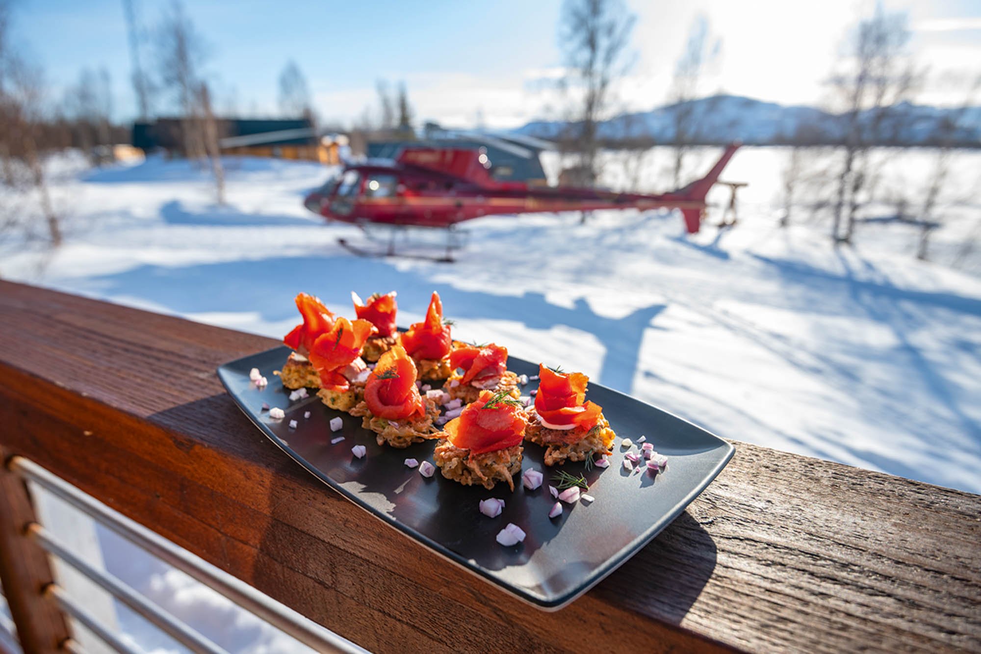 Plate of appetizers with tomato toppings on a patio table outdoors in snowy landscape, helicopter, trees, and mountains in background.