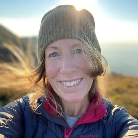 Woman smiling outdoors wearing a gray beanie and a navy jacket with a red collar, with wind blowing her hair.