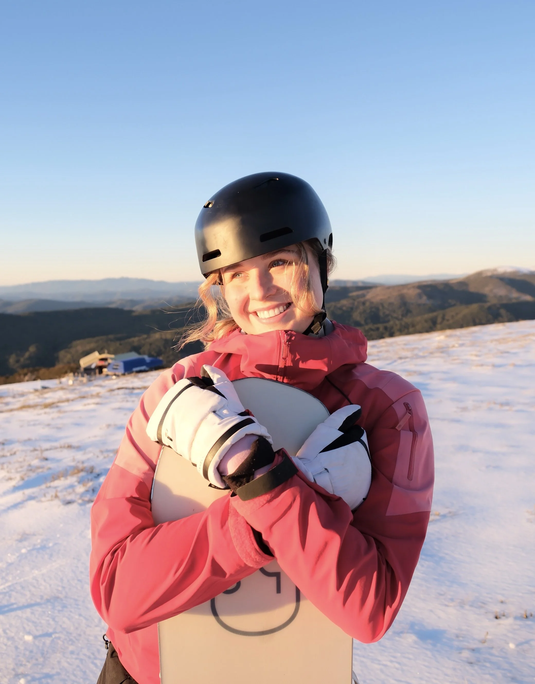 Woman in pink jacket and black helmet holding a snowboard on snowy mountain landscape at sunset.
