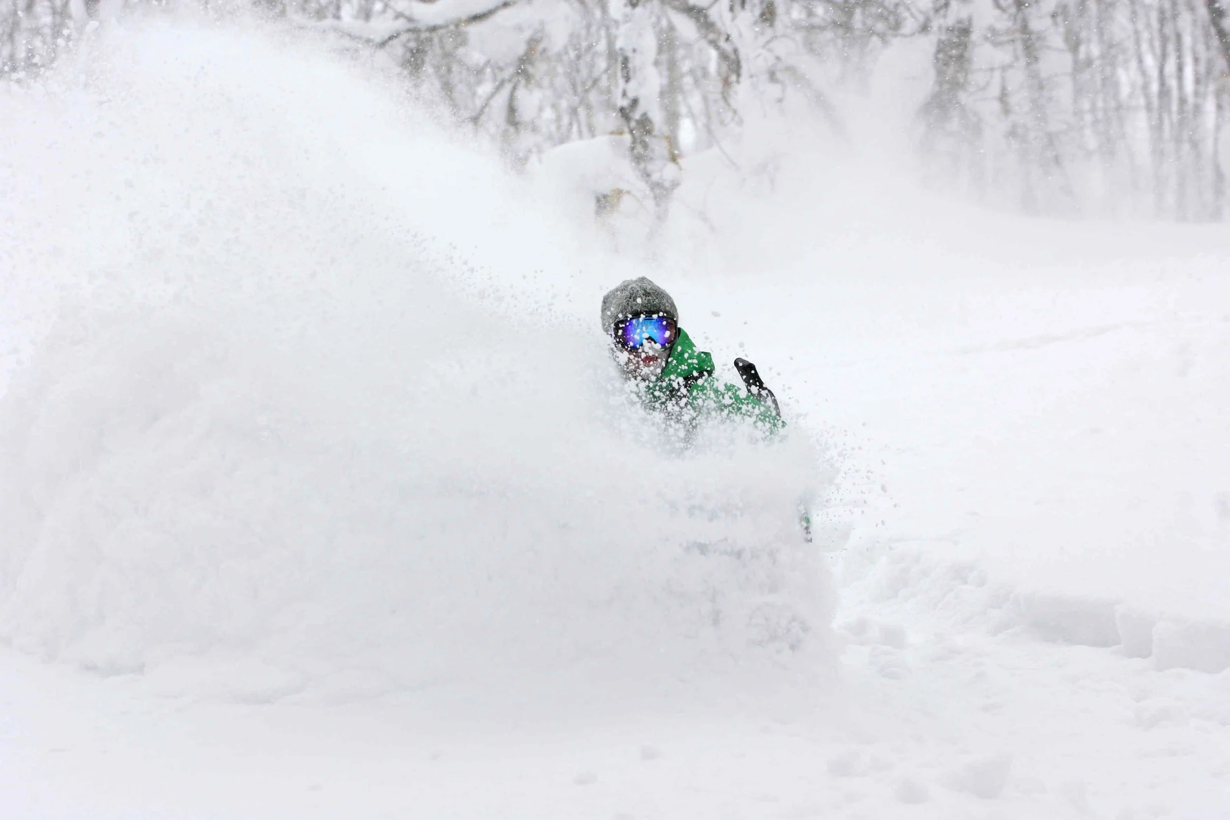 A person wearing a green jacket, helmet, and ski goggles is skiing or snowboarding in deep snow, with snow and trees in the background.
