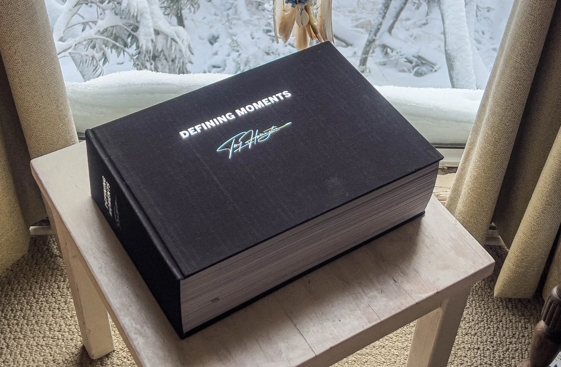 A large black hardcover book titled 'Defining Moments' with a signature font on the cover, resting on a light wooden table near a snow-covered window and beige curtains.