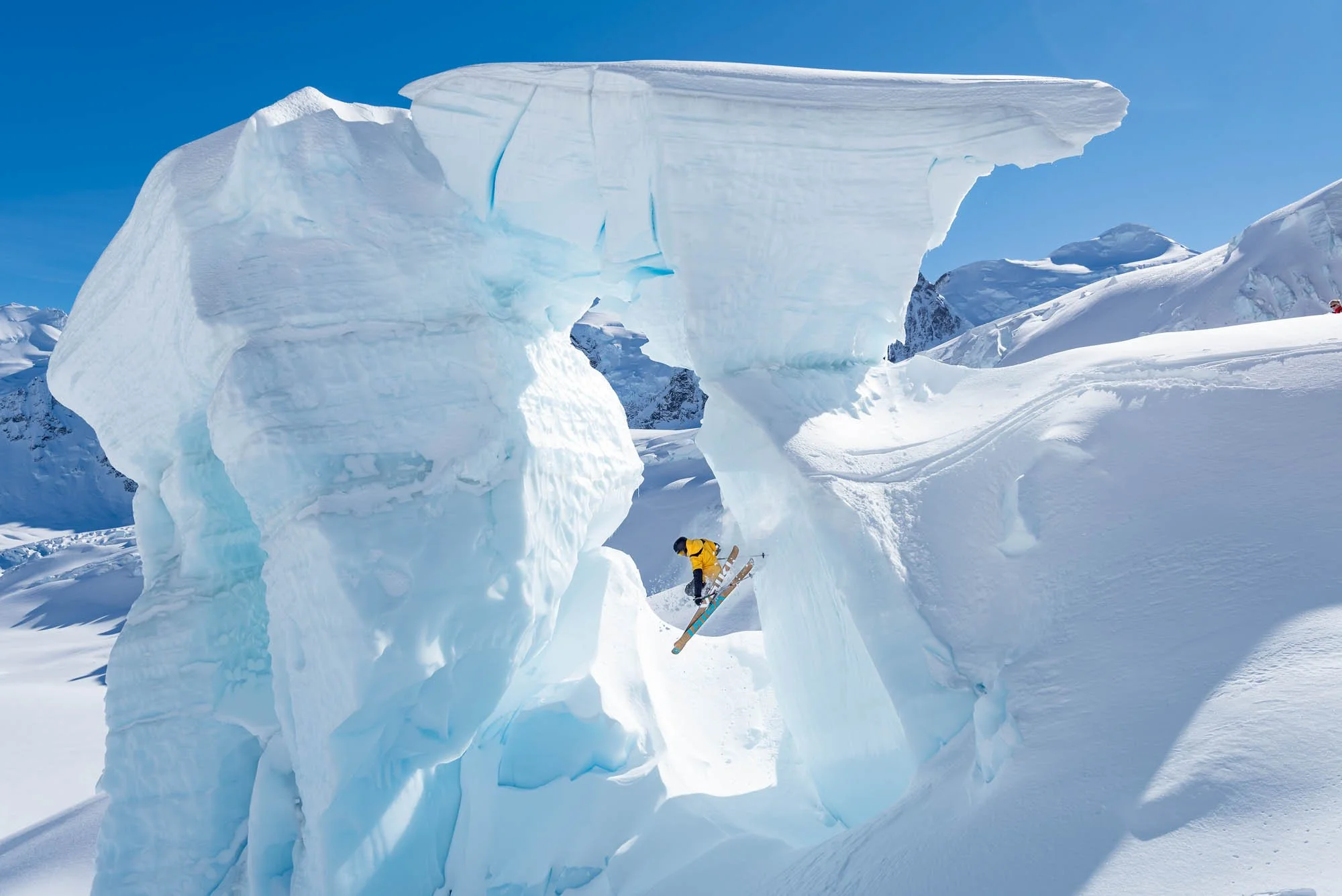 A person in a yellow jacket snowboarding near a large, overhanging ice formation in a snowy, mountainous landscape under a clear blue sky.