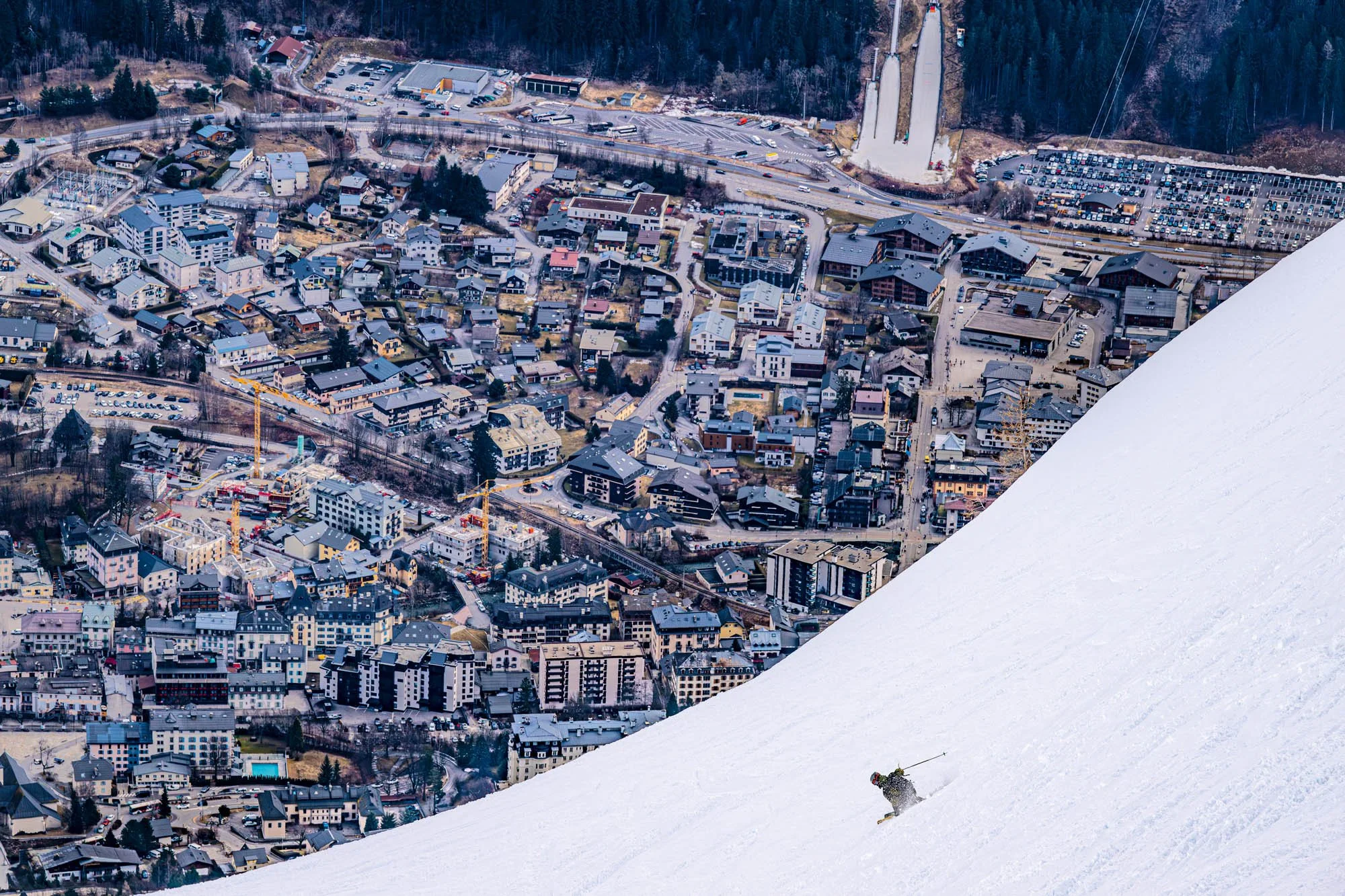 Aerial view of a snow-covered mountain with a skier in the lower right corner and a town with various buildings, roads, and parking lots in the background.