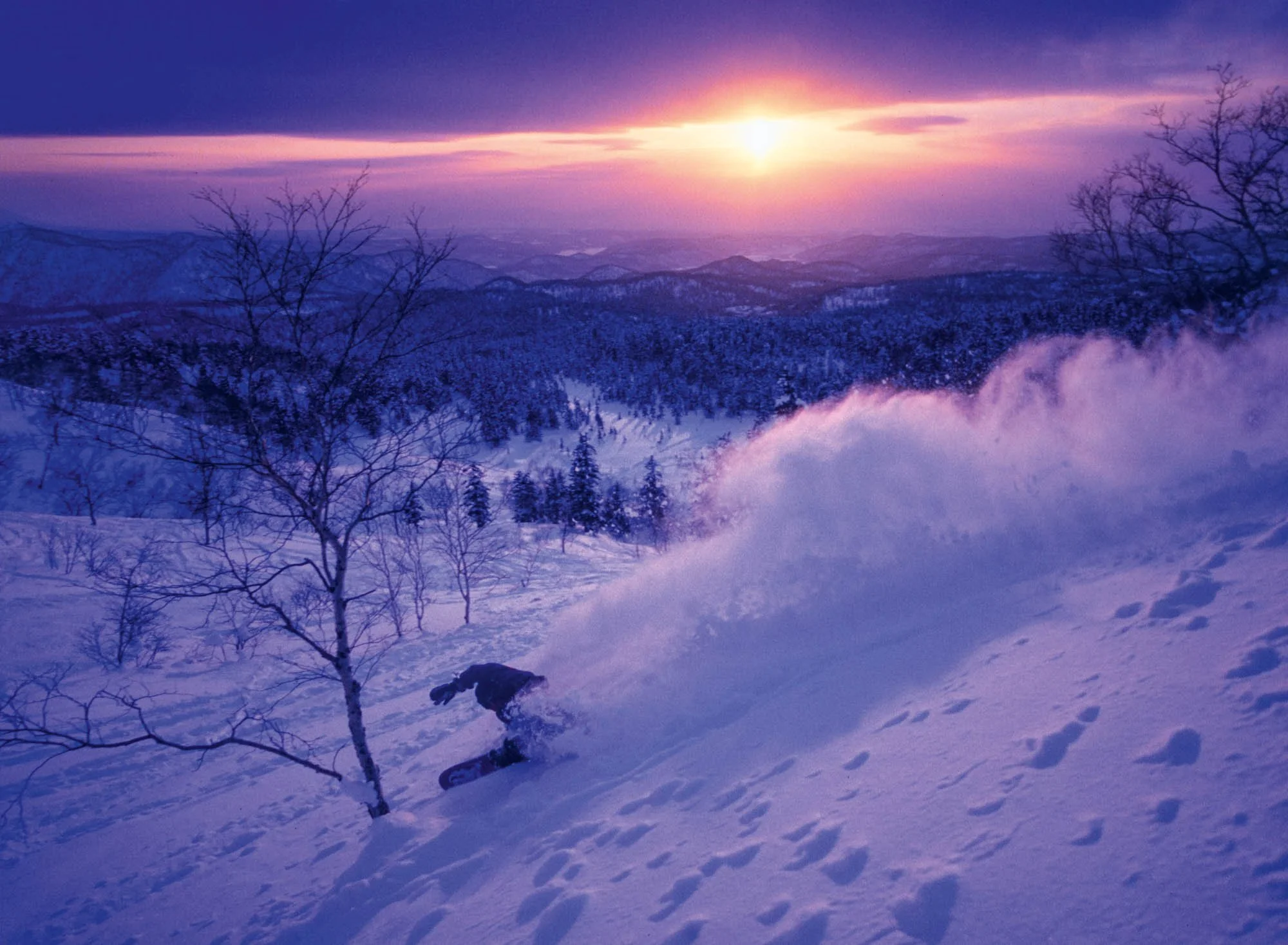 Snow-covered mountain landscape during sunset with leafless trees in the foreground and a ski trail with a skier skiing skiing down the slope.