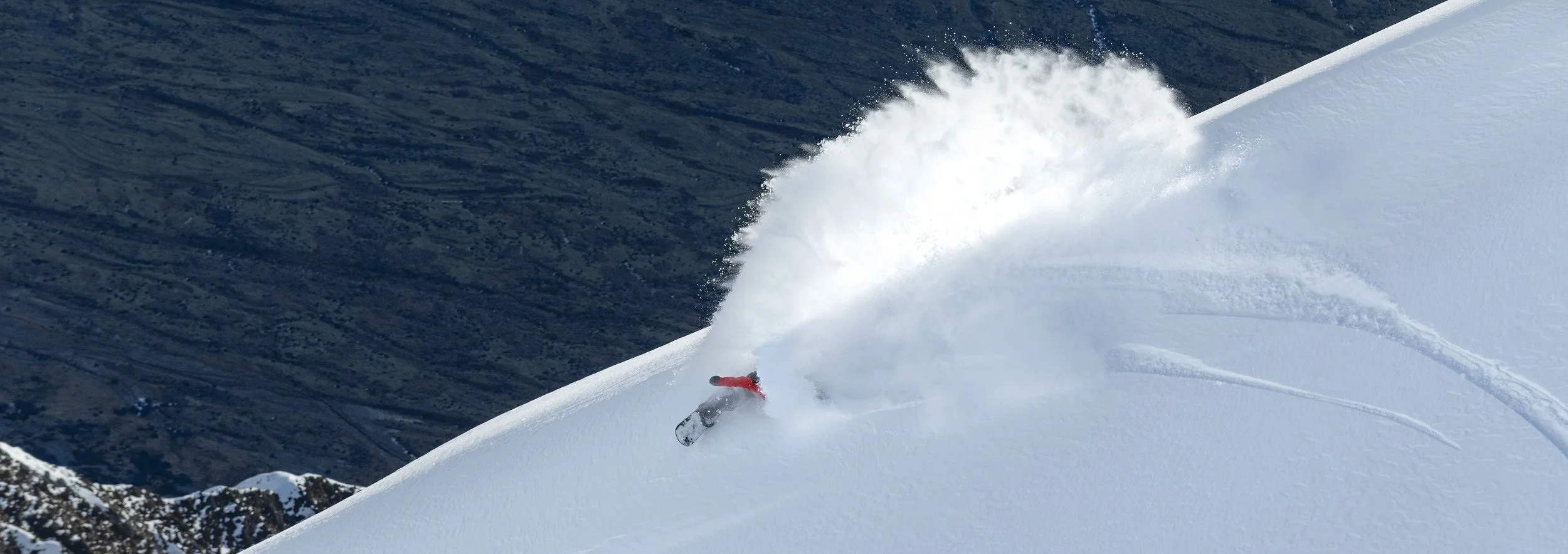 A snowboarder wearing a red jacket is descending a snowy mountain slope, creating a spray of snow as they make a turn.