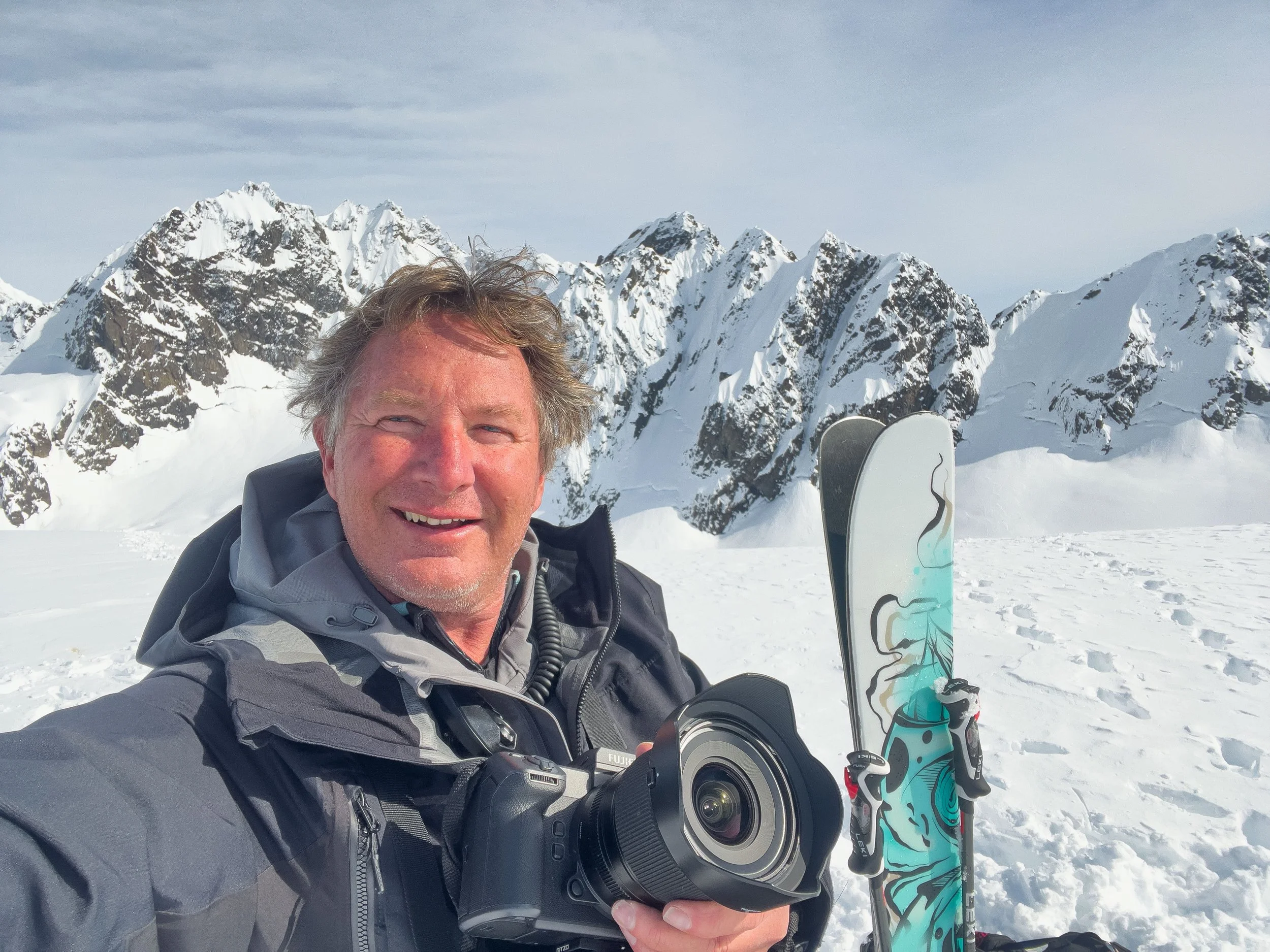 A man taking a selfie in a snowy mountain landscape with snow-covered peaks in the background, holding a snowboard.