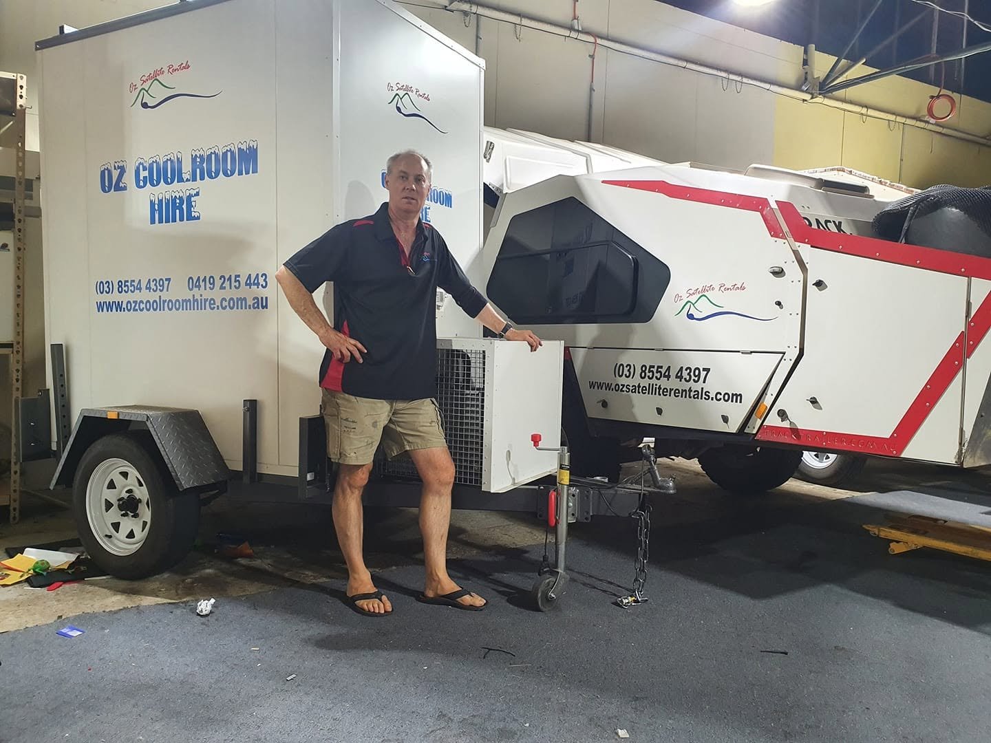 A man standing next to a mobile air conditioning unit on a trailer, with a white and red, futuristic-looking vehicle in the background. The man is wearing a black and red shirt, khaki shorts, and flip flops.